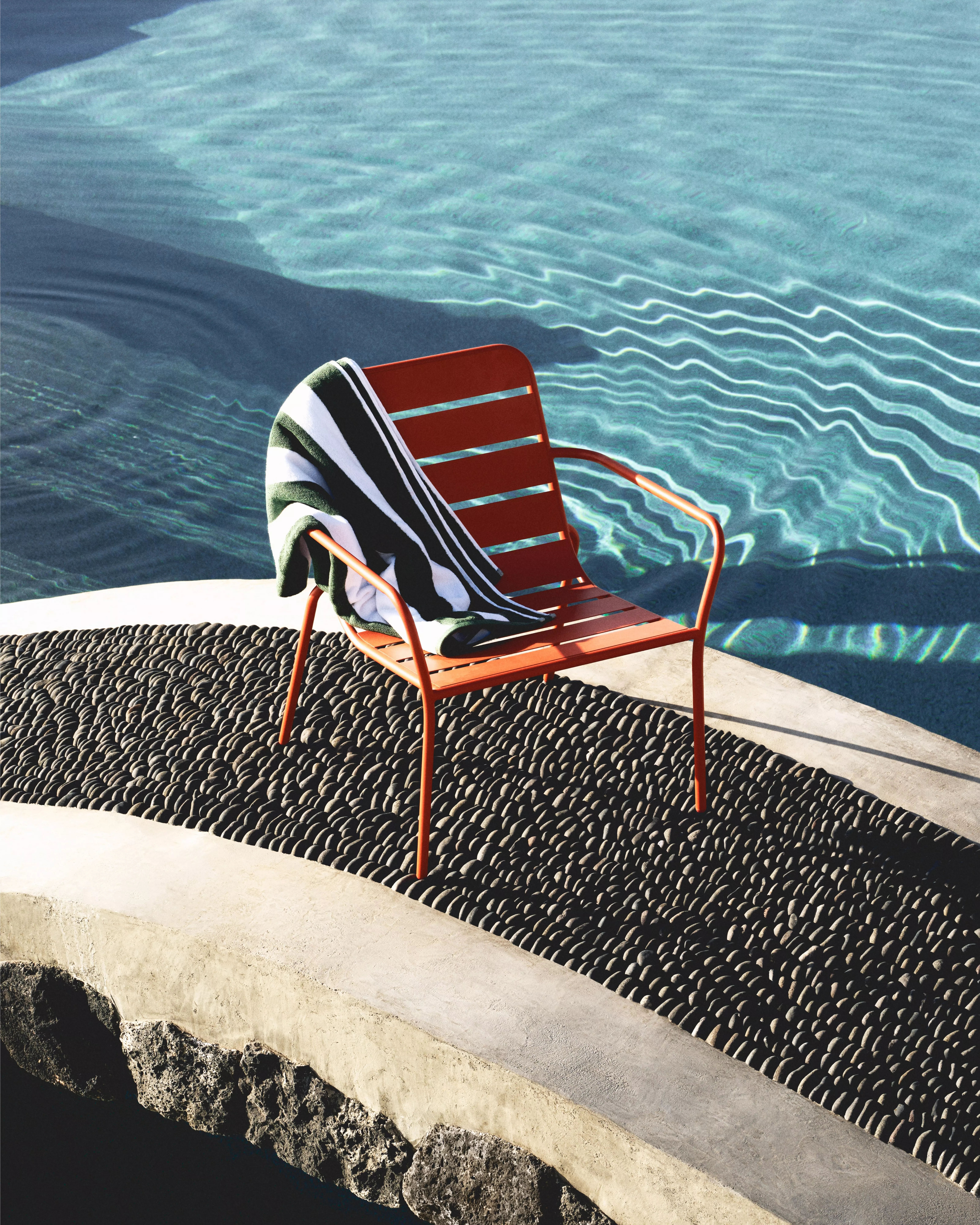 A red chair with a striped towel sits on a stone path by a calm pool, casting reflections in the water.