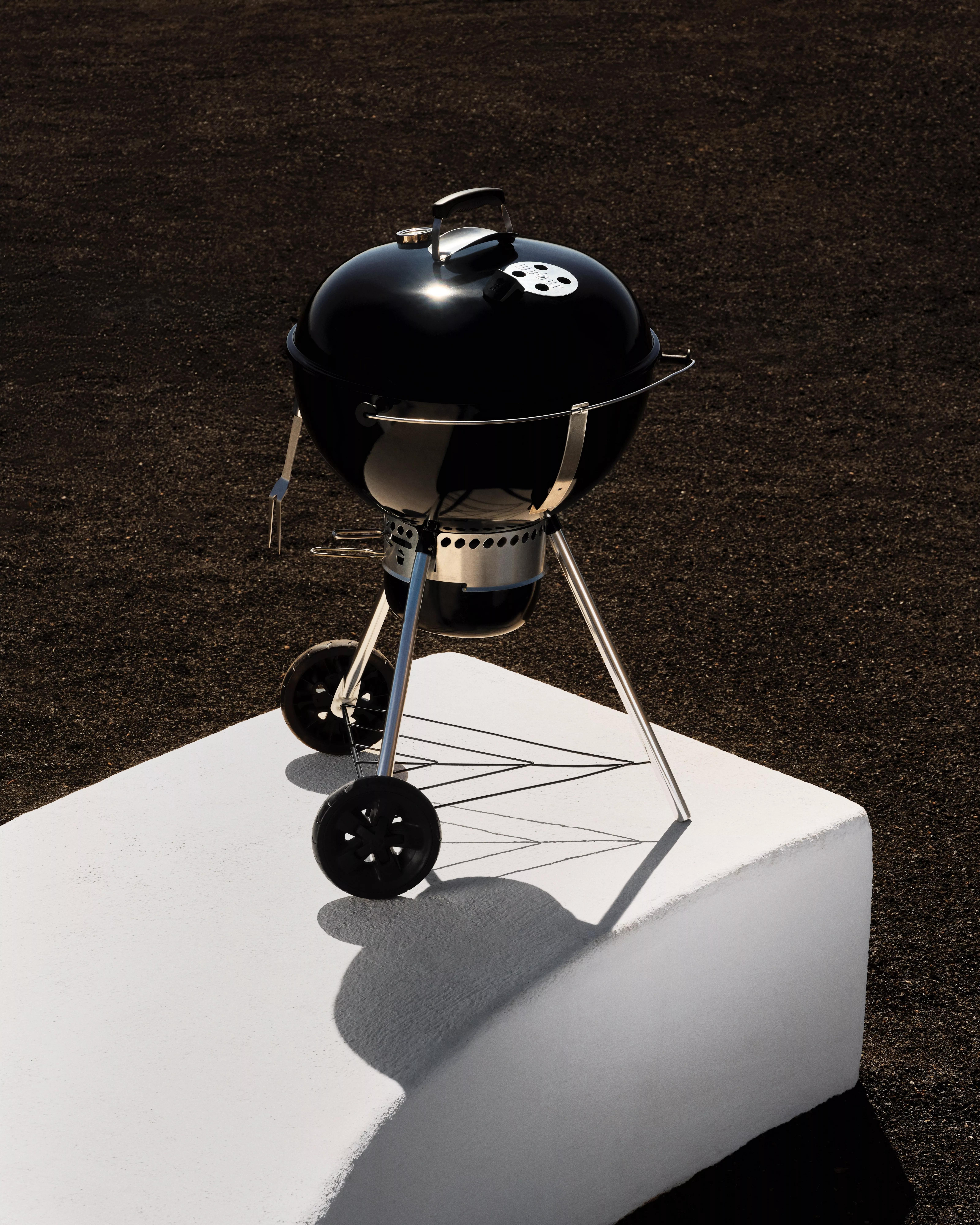 A black kettle barbecue grill stands on a white platform against a dark, textured background.