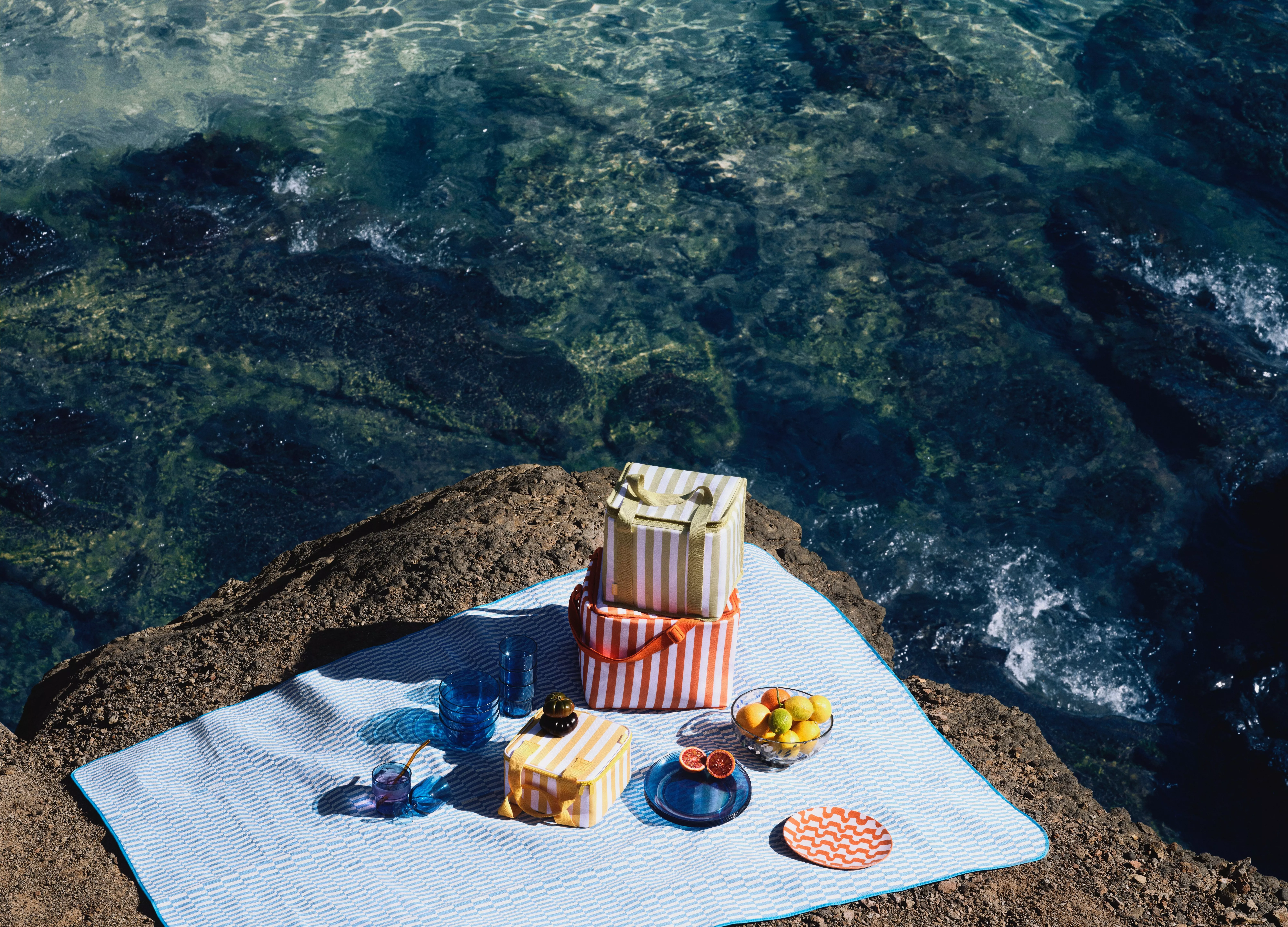 Colourful picnic setup on a rocky seaside. Striped coolers, vibrant plates, and fresh fruit enhance the scene.