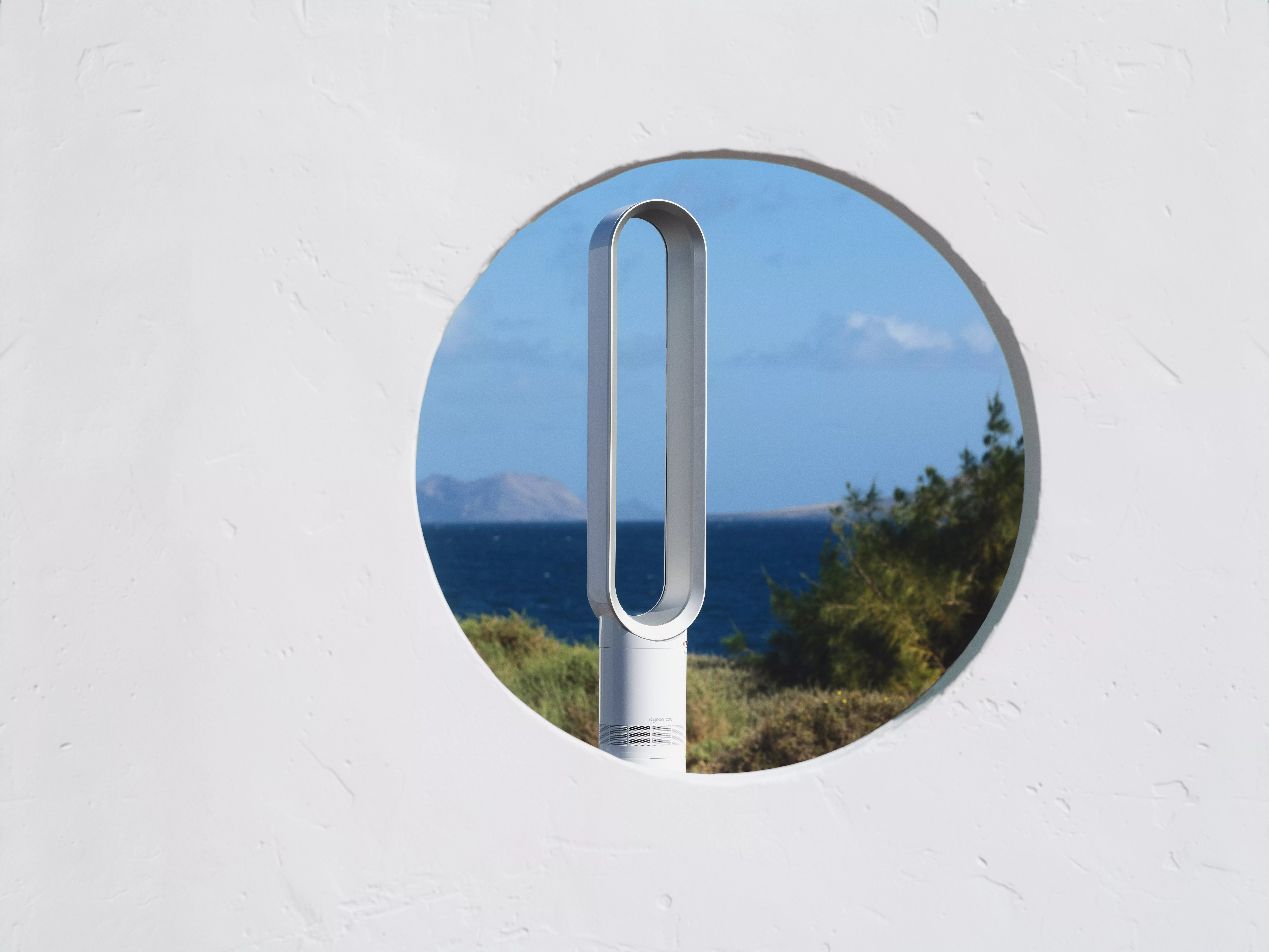 Modern bladeless fan framed by a circular window, with a scenic ocean view and greenery in the background.