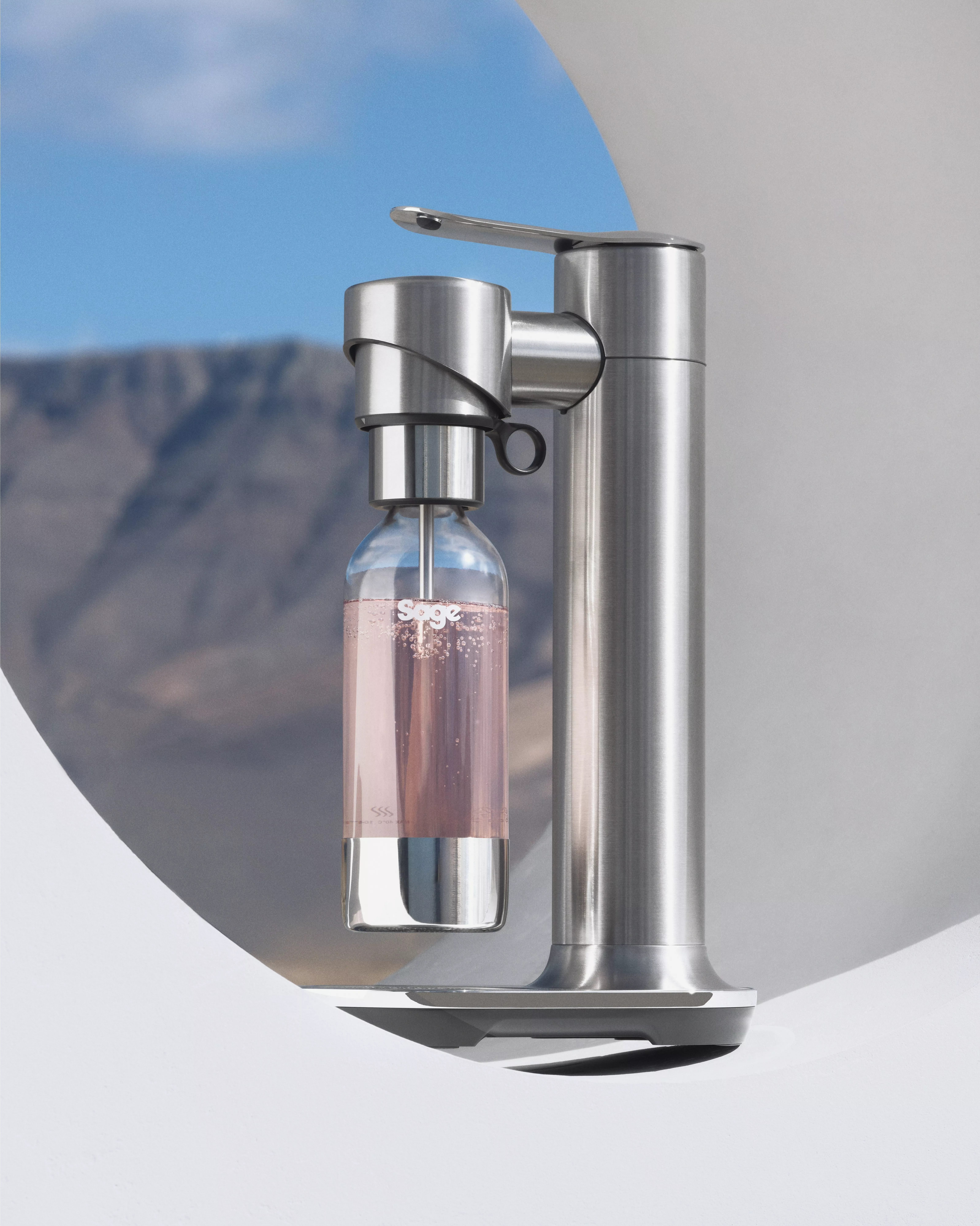 Sleek silver soda maker with a bottle of pink fizzy drink, set against a mountainous outdoor backdrop.