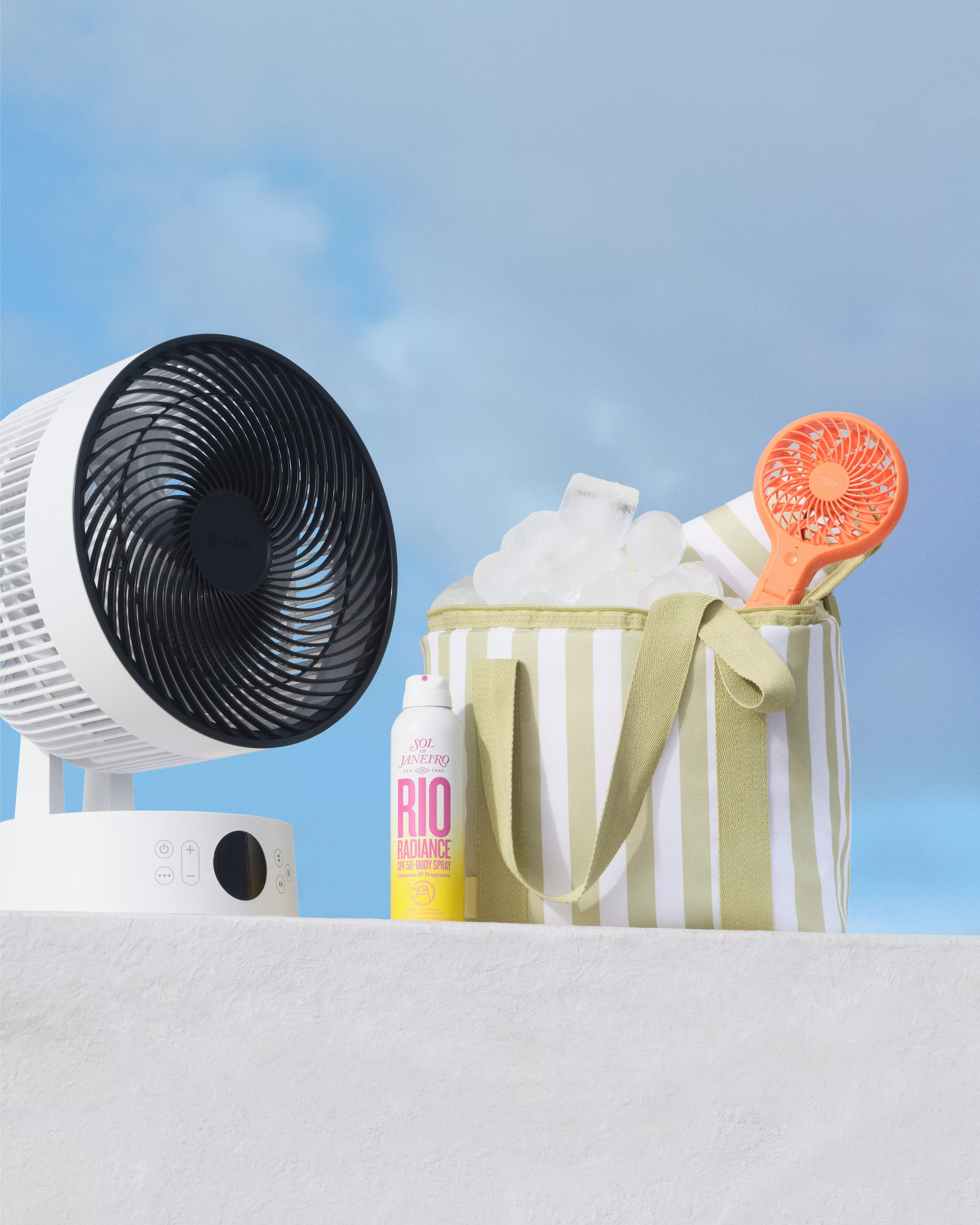 A white fan, striped cooler bag with ice, orange handheld fan, and sunscreen under a blue sky.