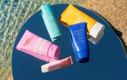 Variety of sunscreen tubes on a reflective surface, near water and sand, showcasing sun protection products.