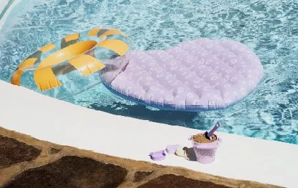 Inflatable rings float in a sunlit pool, with a sand-filled toy bucket and spade on the stone poolside.