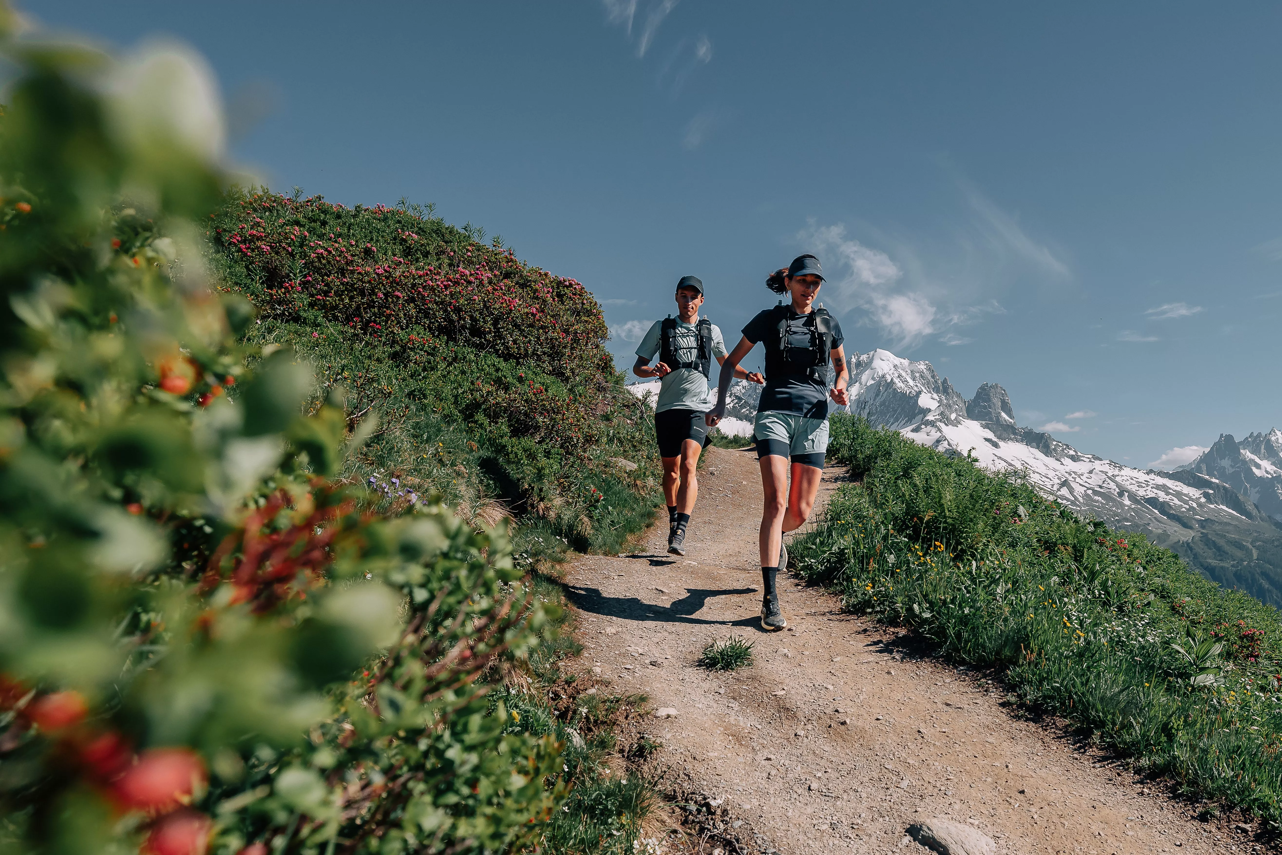 Two trail runners on a mountain path with backpacks, wildflowers and snow-capped peaks under blue sky
