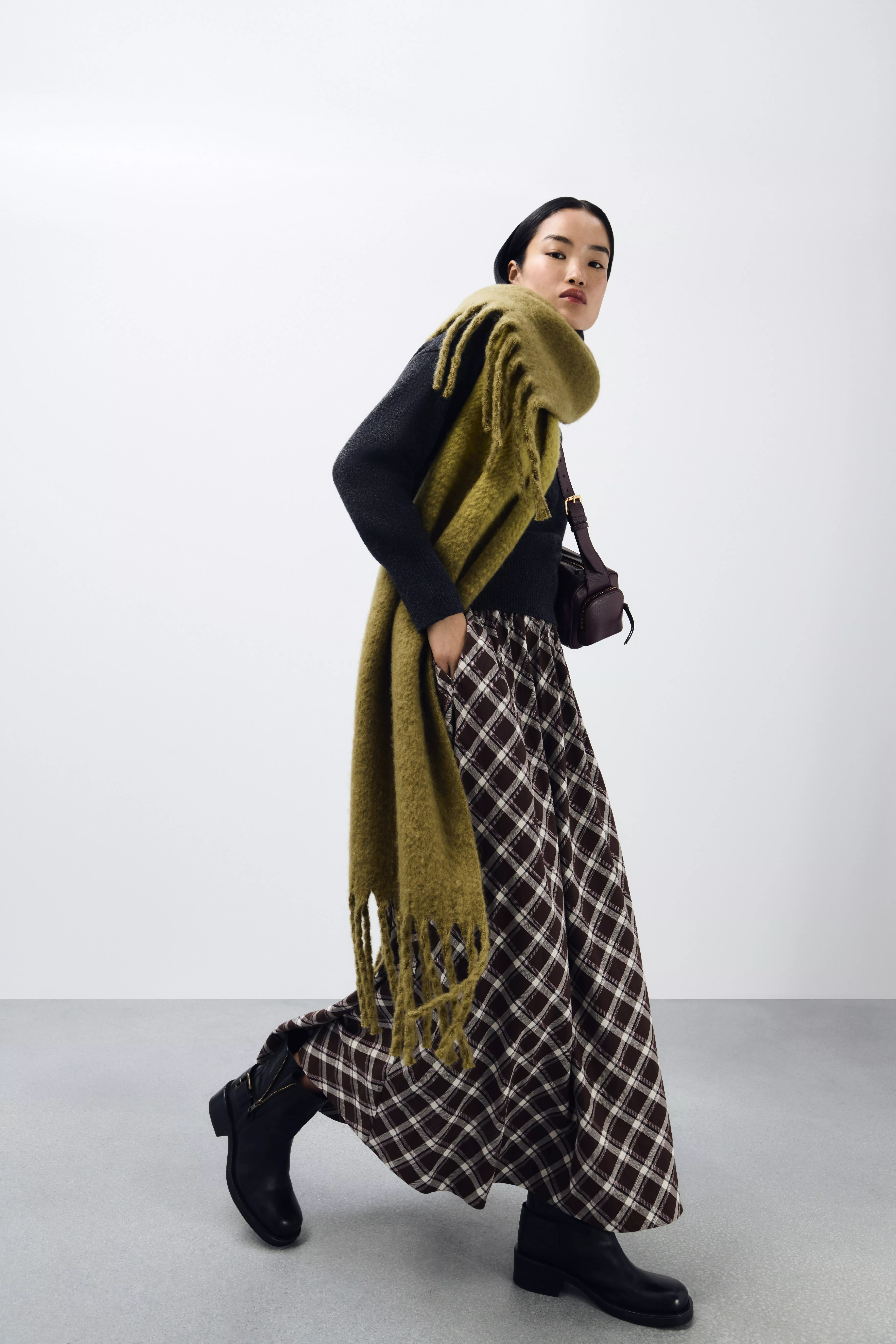 A fashion model wearing a black top, a long, fringed olive-green shawl, and a checkered brown and black skirt.