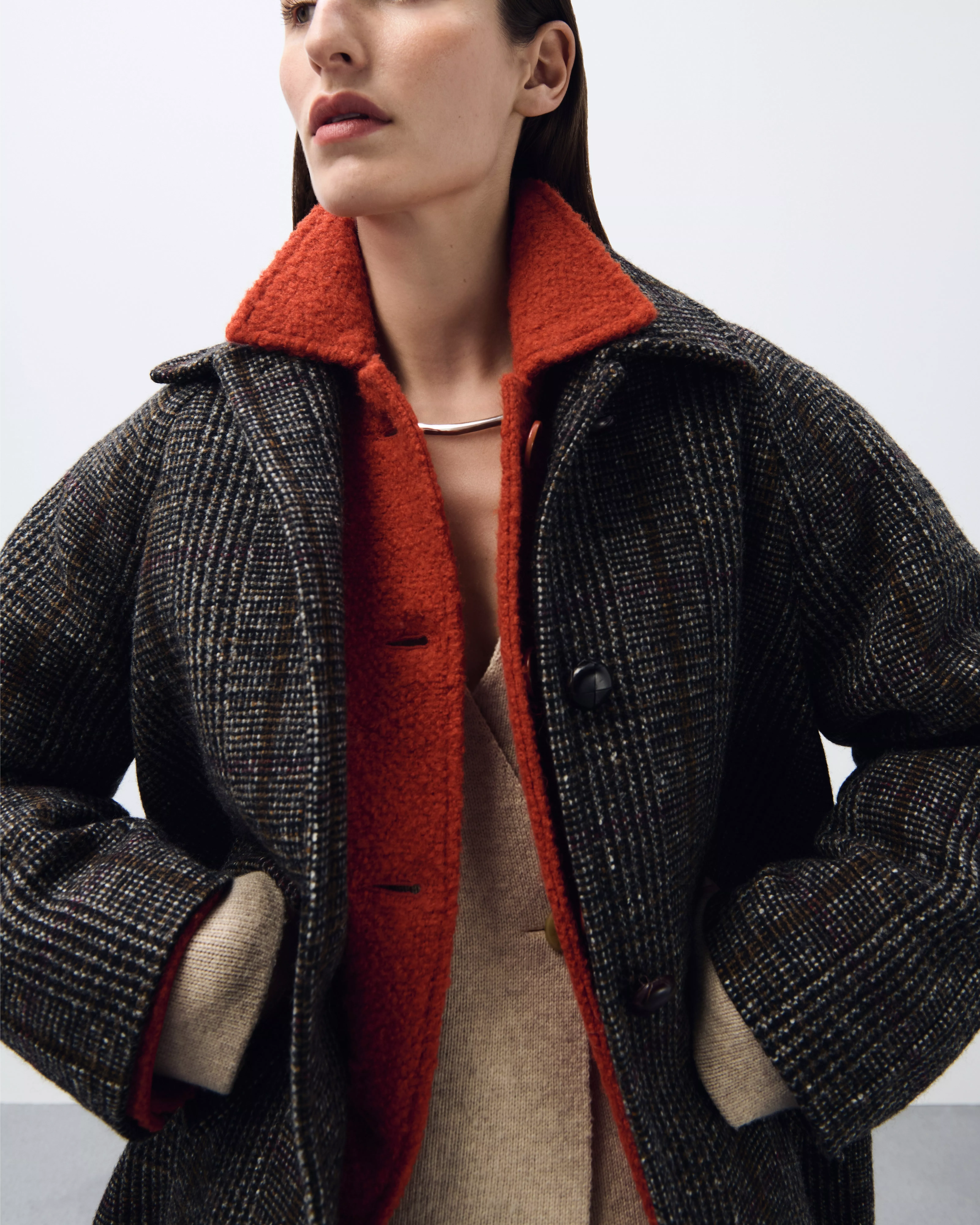 A close-up shot of a woman wearing a dark grey, tweed-like coat with an exposed vibrant orange/red collar lining and a silver necklace.