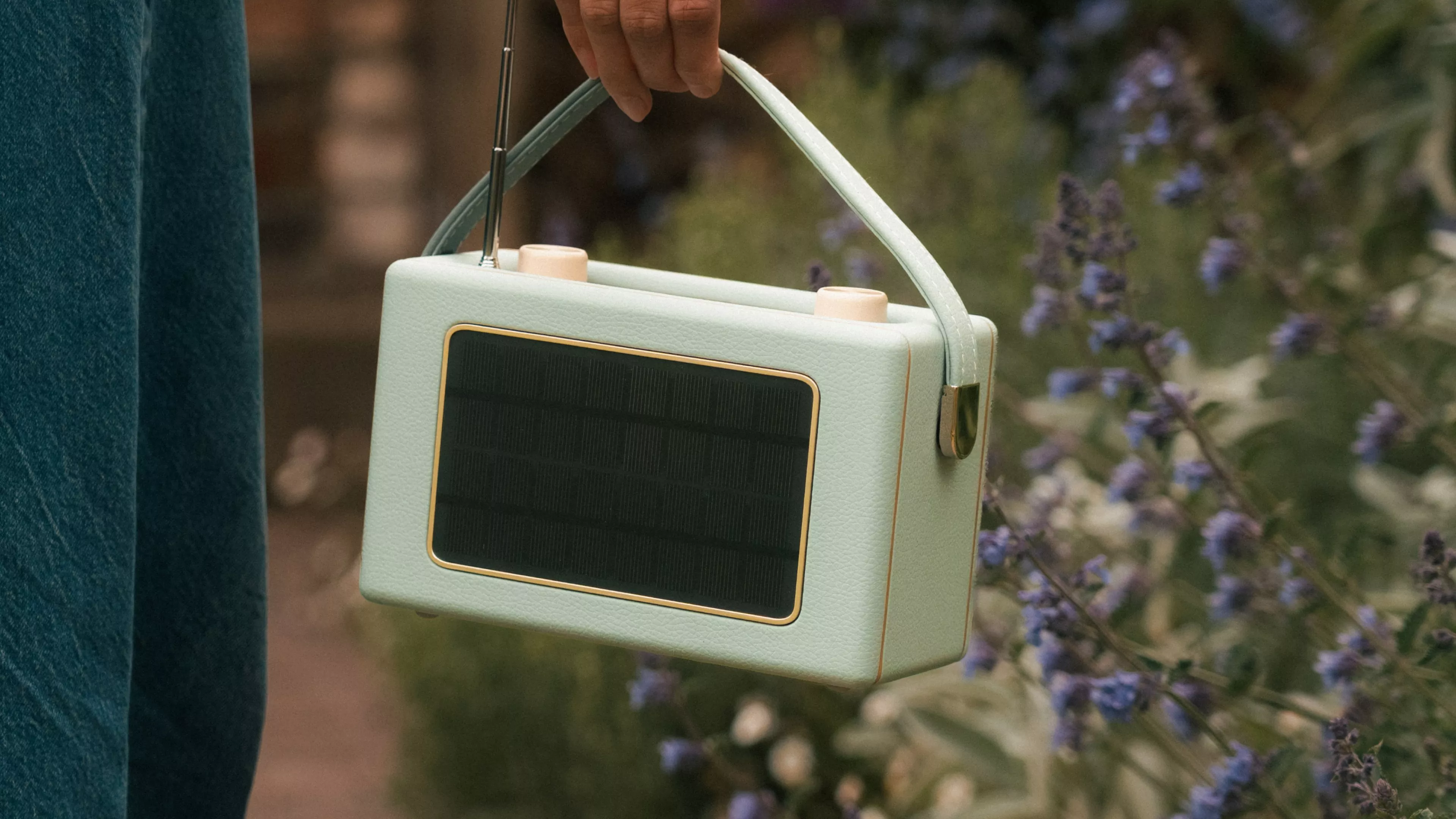 Hand holding mint green solar powered portable radio with antenna, outdoors beside purple garden flowers