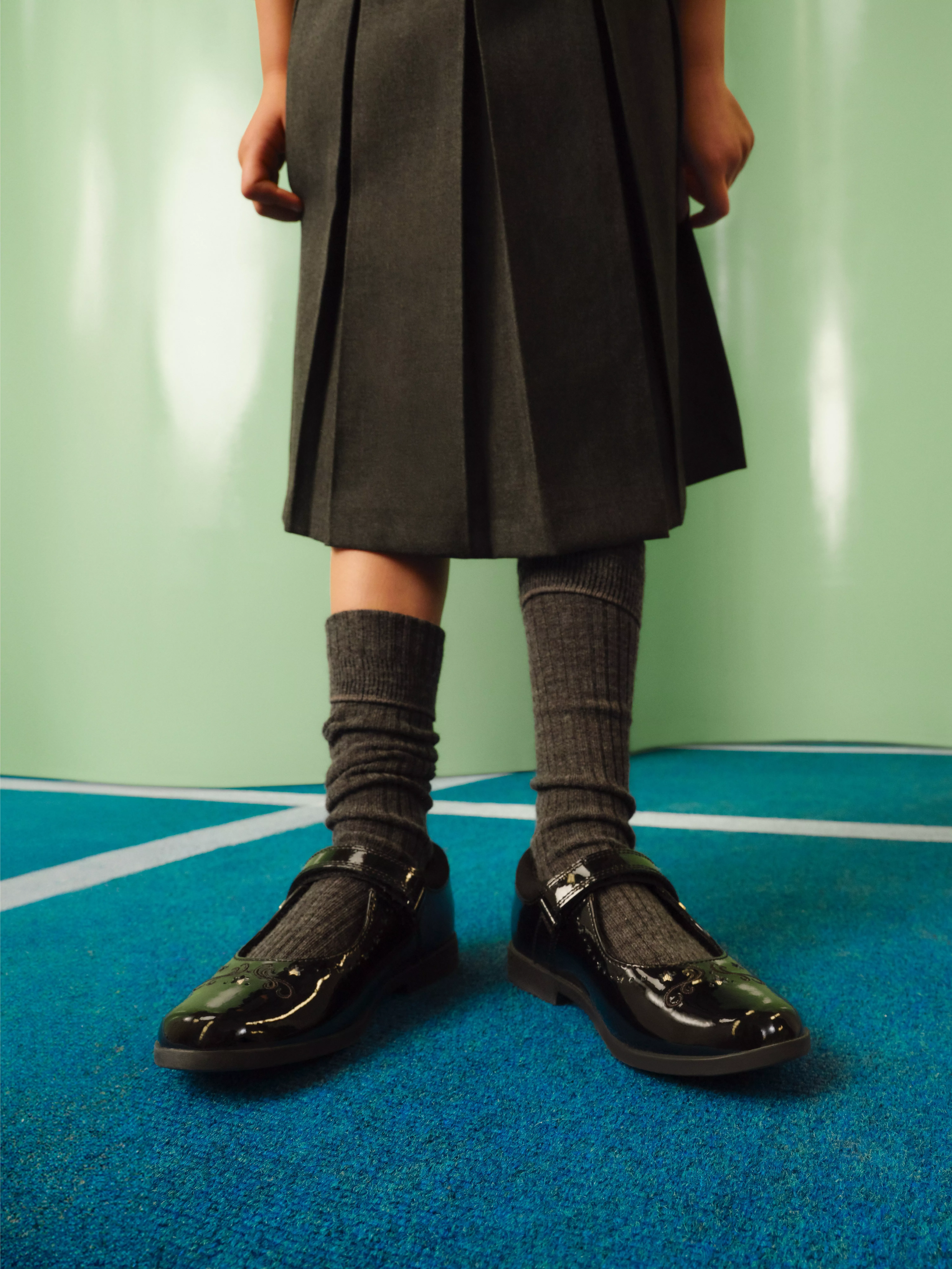 Child wearing grey school uniform skirt, knee-high socks and black patent shoes standing on blue sports court.