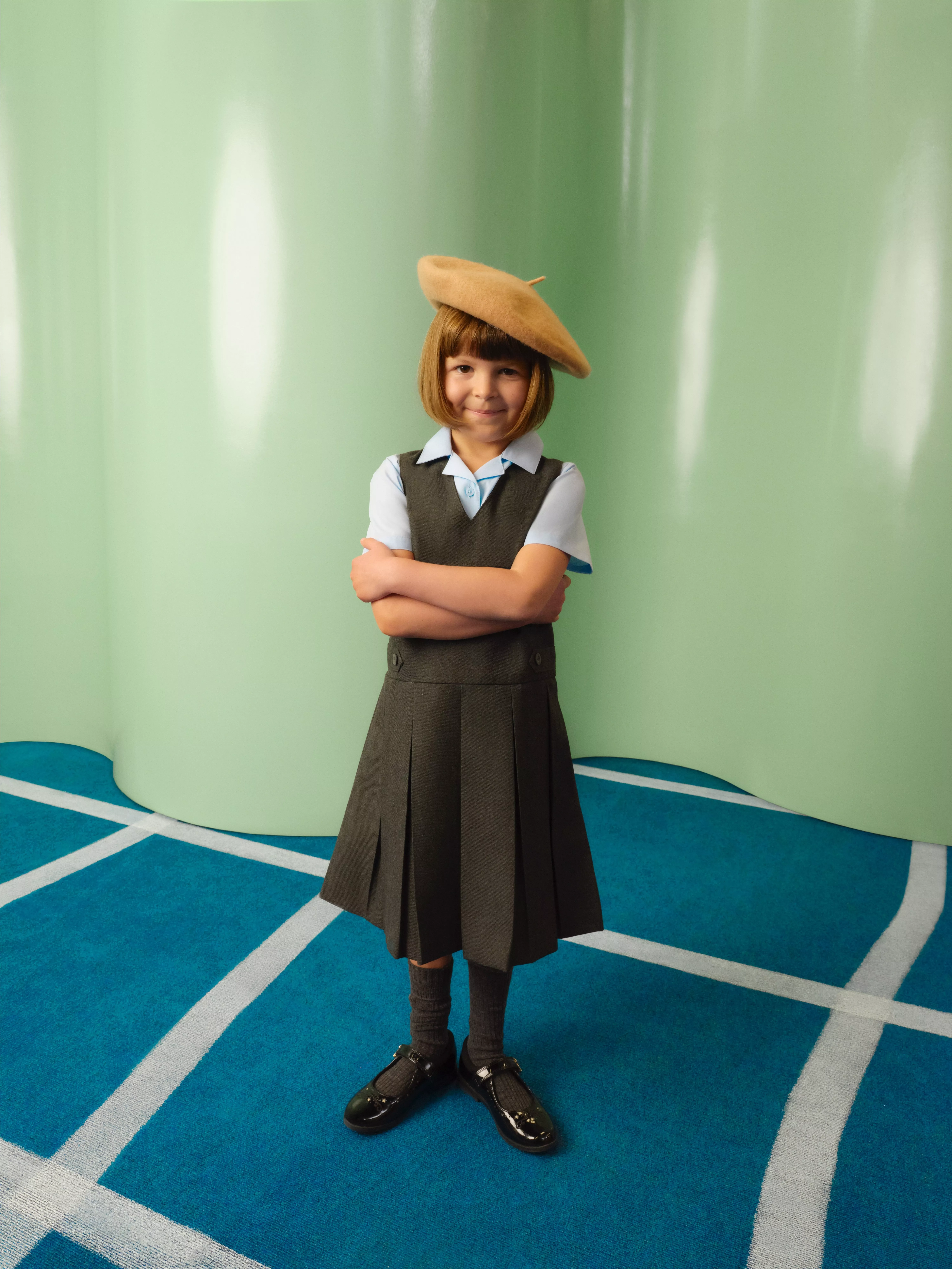 Smiling schoolgirl in grey uniform and beret, arms crossed, standing on blue carpet with green backdrop