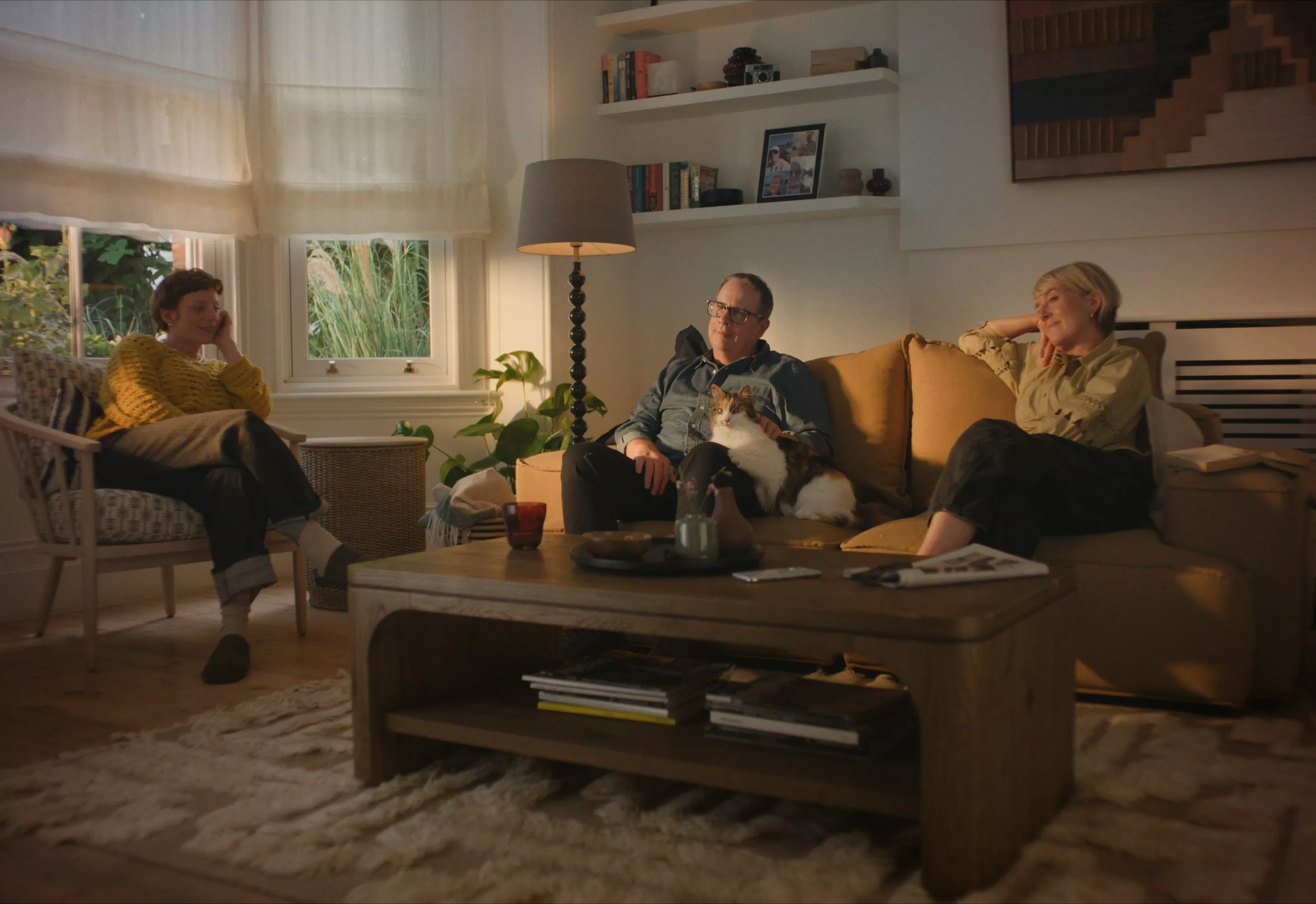 Cosy family living room with three people on sofas and armchair, cat resting beside man, warm light