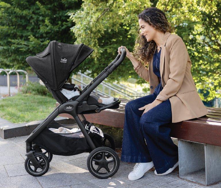 A woman in a tan blazer sits on a park bench, smiling at a baby in a black Joie stroller. Lush green trees and a paved path are in the background of the bright, outdoor scene.