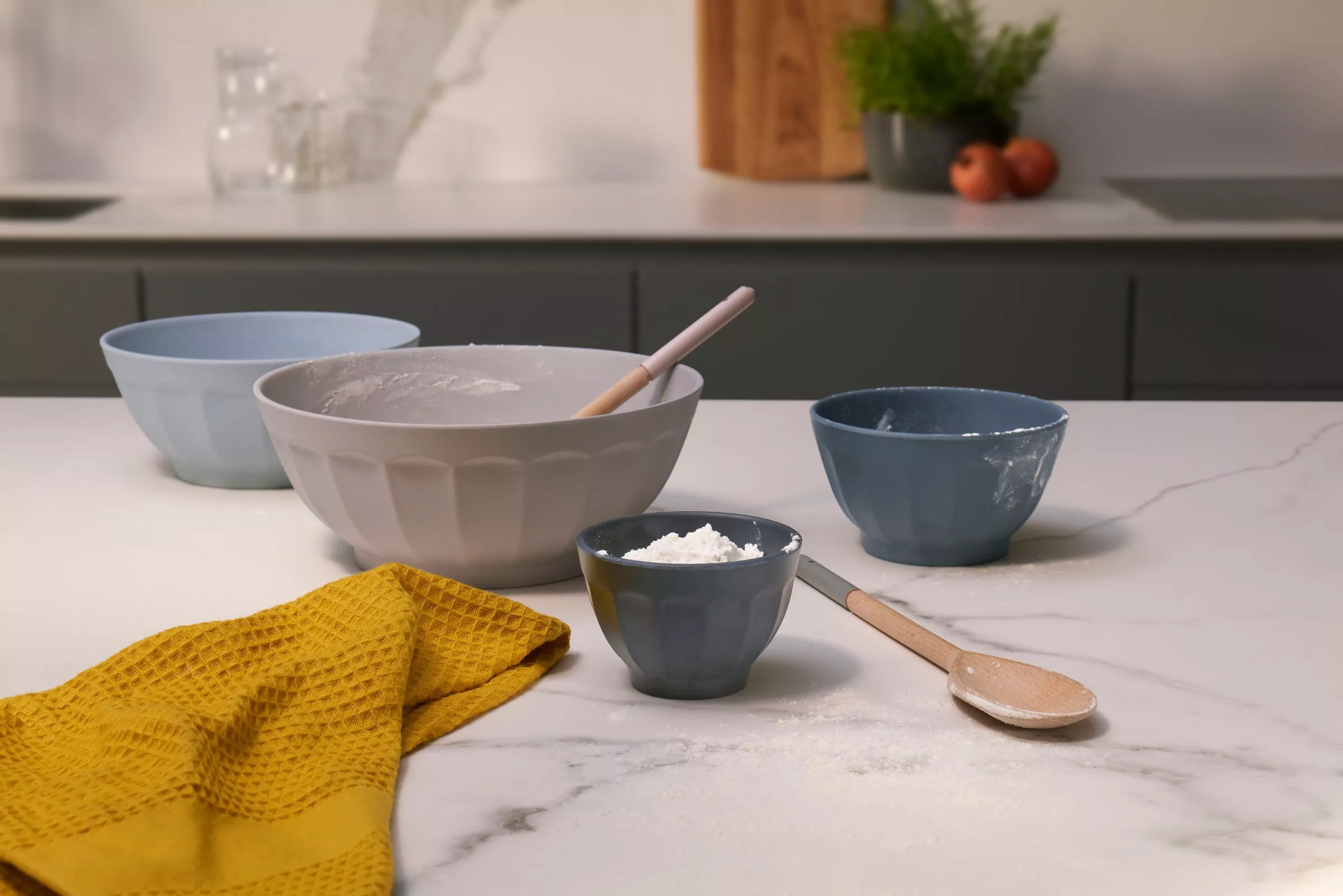 Ceramic mixing bowls with flour, wooden spoon and yellow tea towel on marble kitchen worktop, baking prep scene
