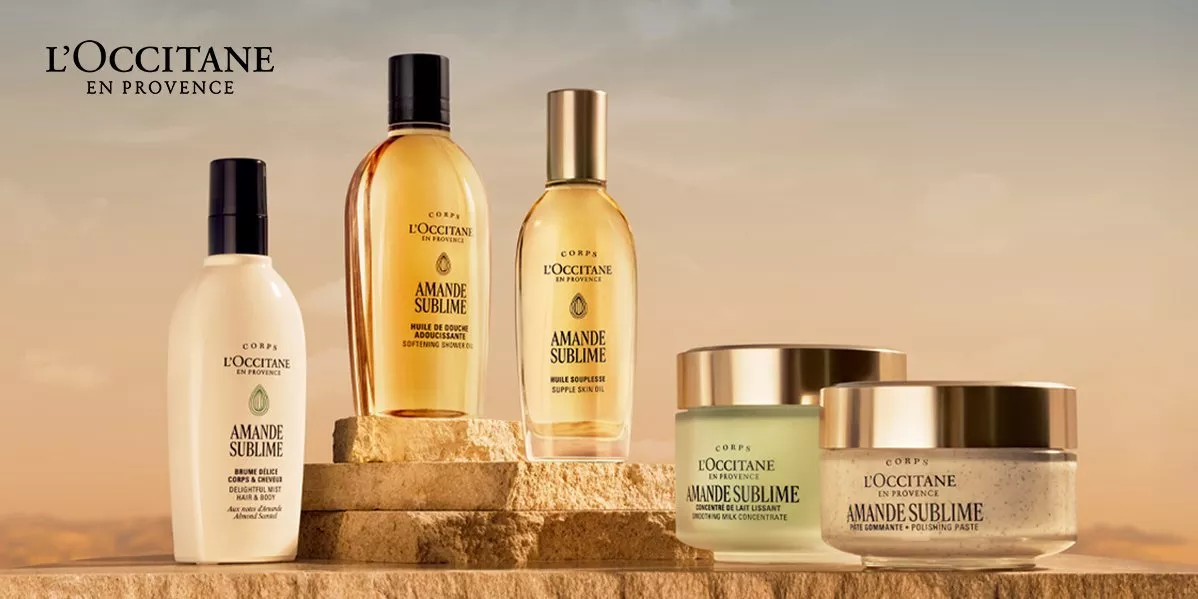 A collection of five L'Occitane Amande Sublime almond-based body care products arranged on tiered sandstone blocks against a warm, sunset-lit background.