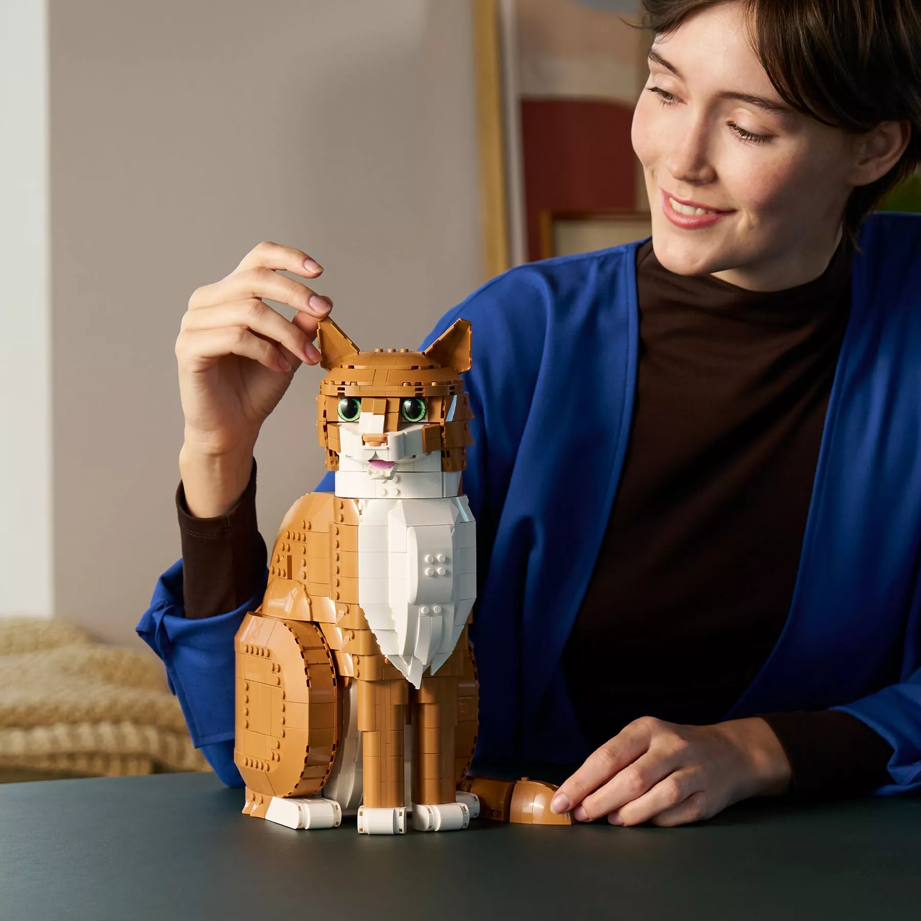 Smiling woman adjusts a large LEGO brick-built ginger and white cat model on a table indoors