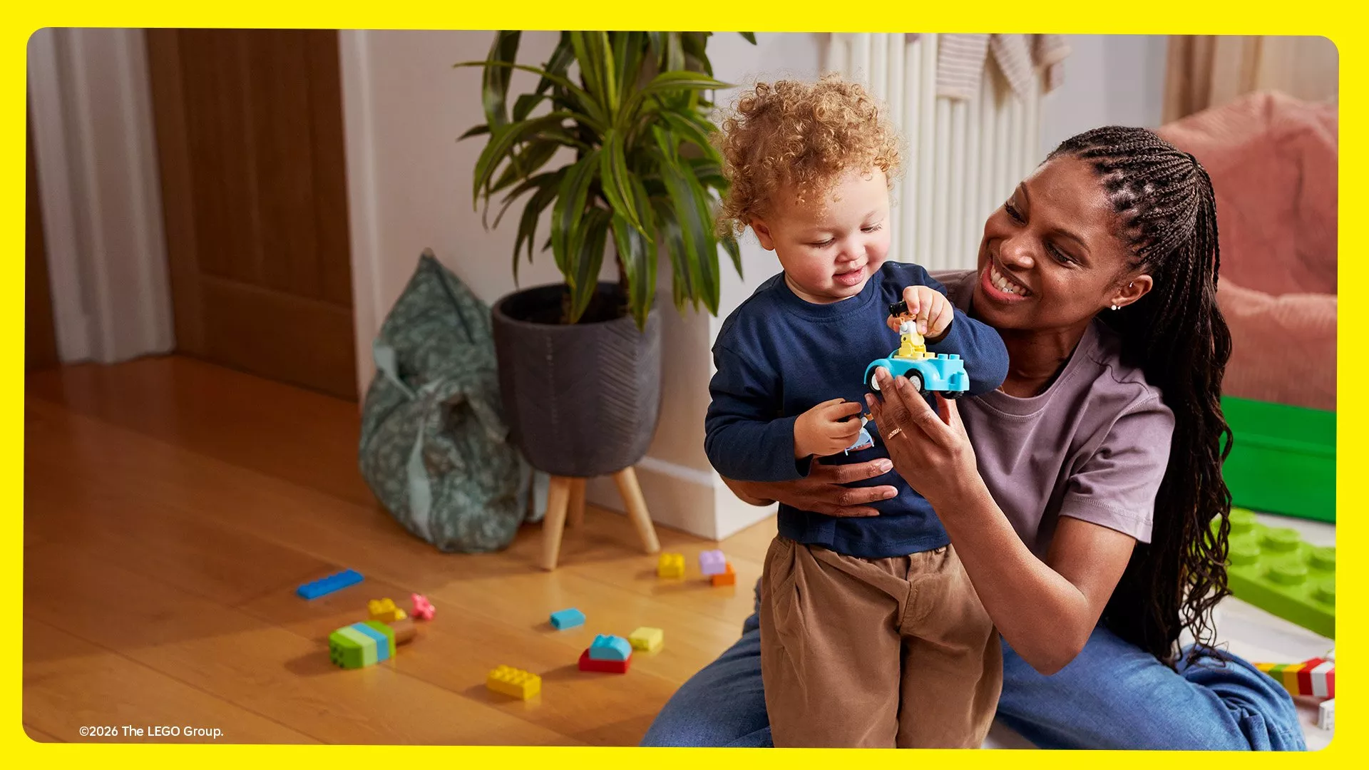 A smiling woman with braided hair sits on the floor assisting a young toddler with curly hair, who is playing with a light blue LEGO Duplo toy car.