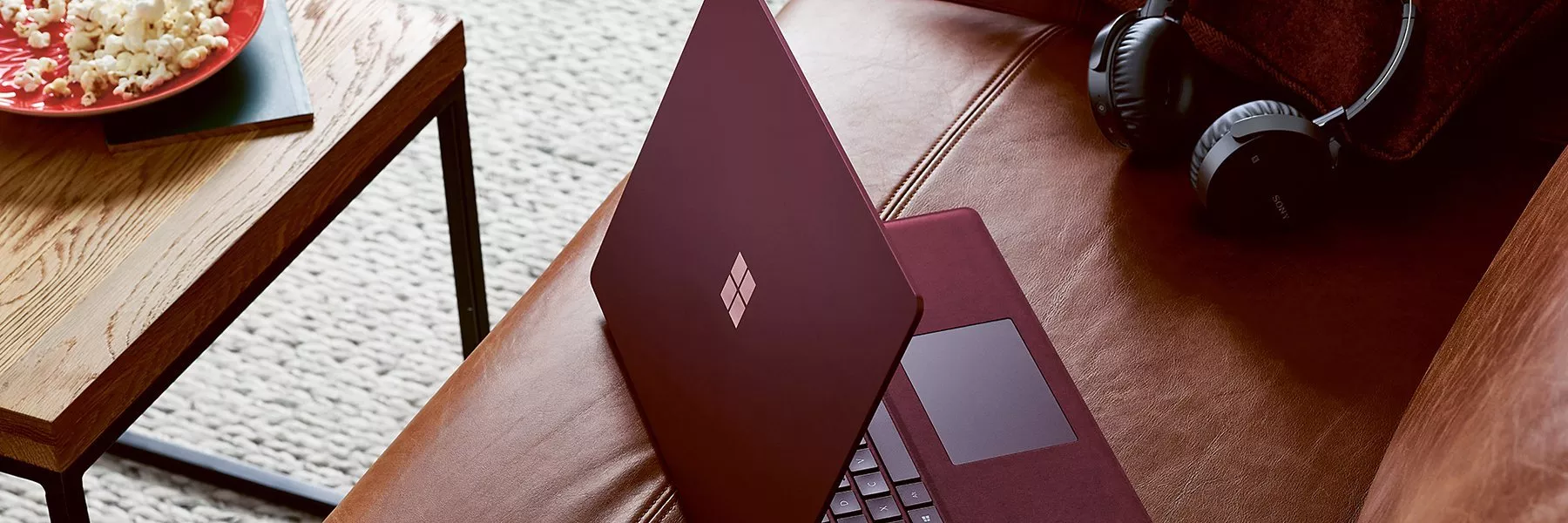 A maroon laptop on a brown leather sofa with headphones nearby; a table with popcorn is in the background.