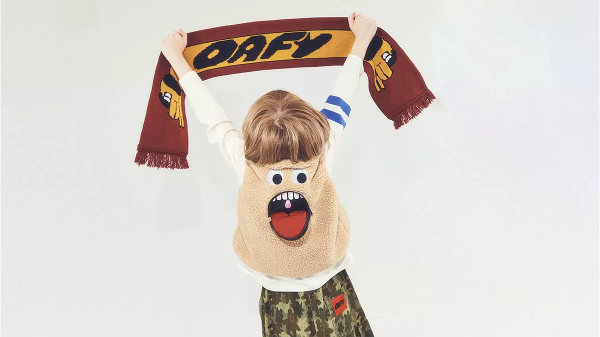 Child holding an OAFY scarf overhead, wearing a fluffy cartoon face gilet and camo trousers on white backdrop