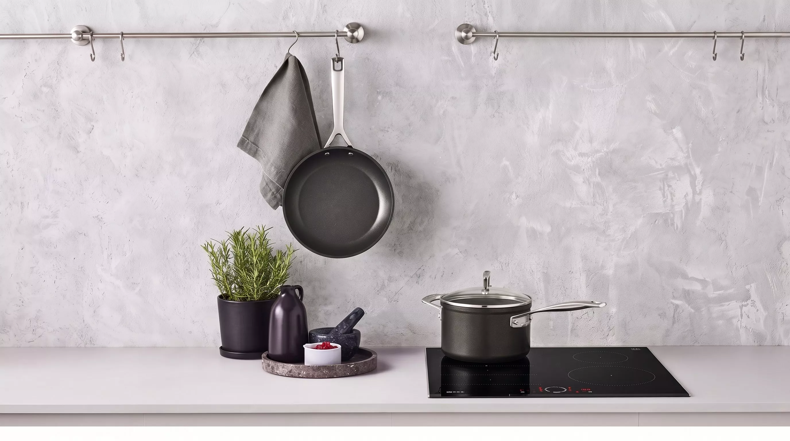 Black non-stick frying pan hanging on a grey wall next to a green potted plant and a black cooking pot on a gas hob.