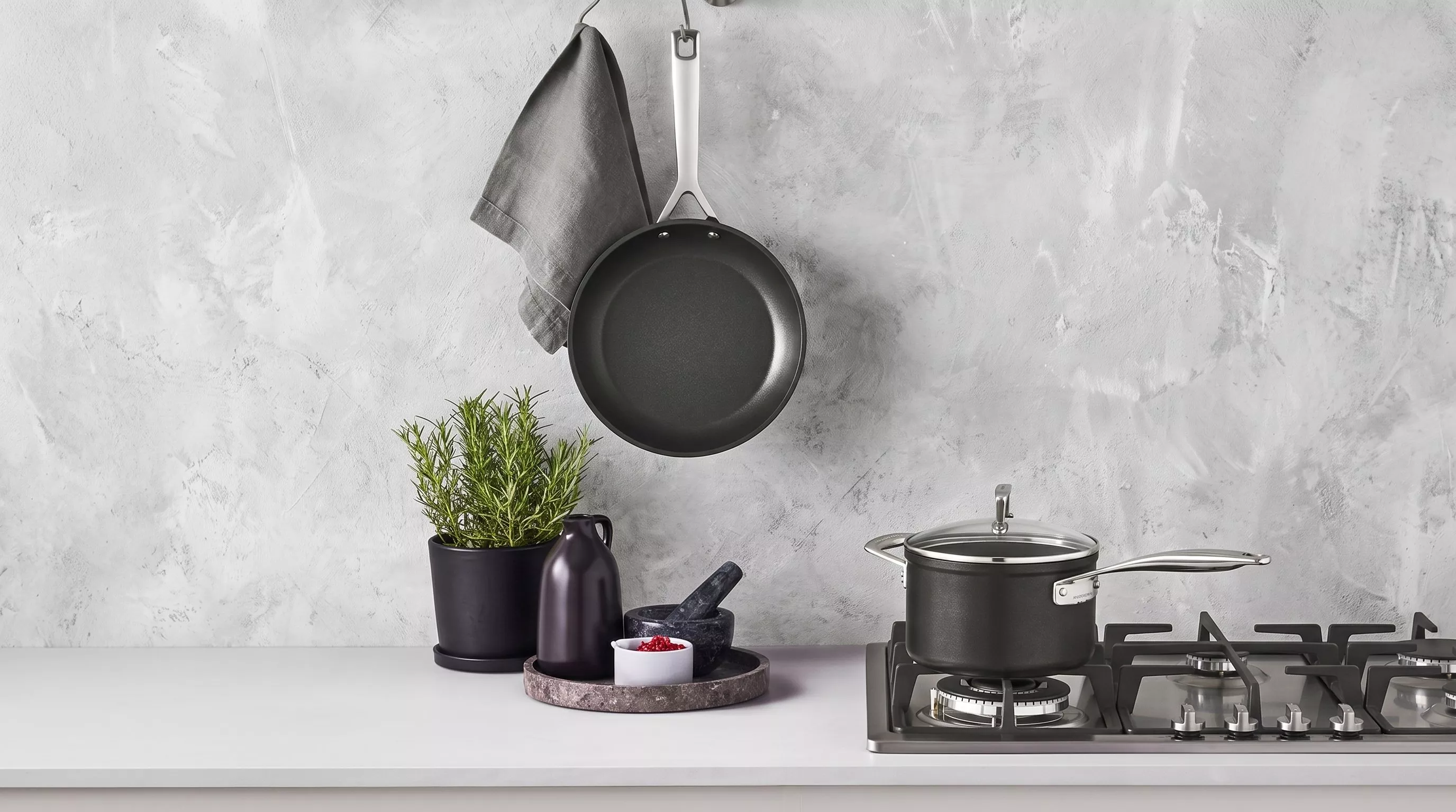 Black non-stick frying pan hanging on a grey wall next to a green potted plant and a black cooking pot on a gas hob.