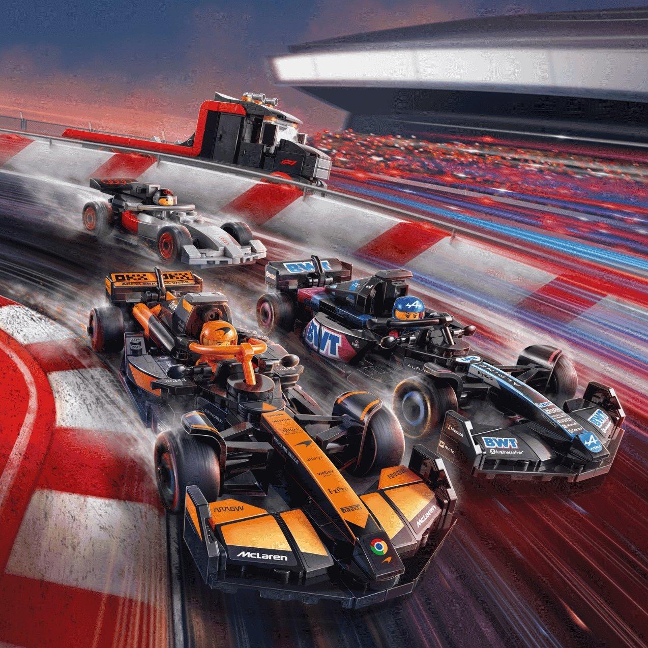 Lego Formula 1 racing cars speed around a racetrack curve with motion blur. A McLaren car leads an Alpine and another F1 car, with a grandstand in the background.