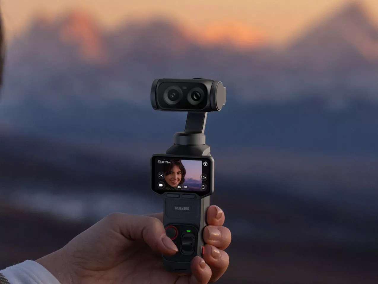 Hand holding Insta360 gimbal action camera filming selfie video, with blurred mountain sunset backdrop outdoors