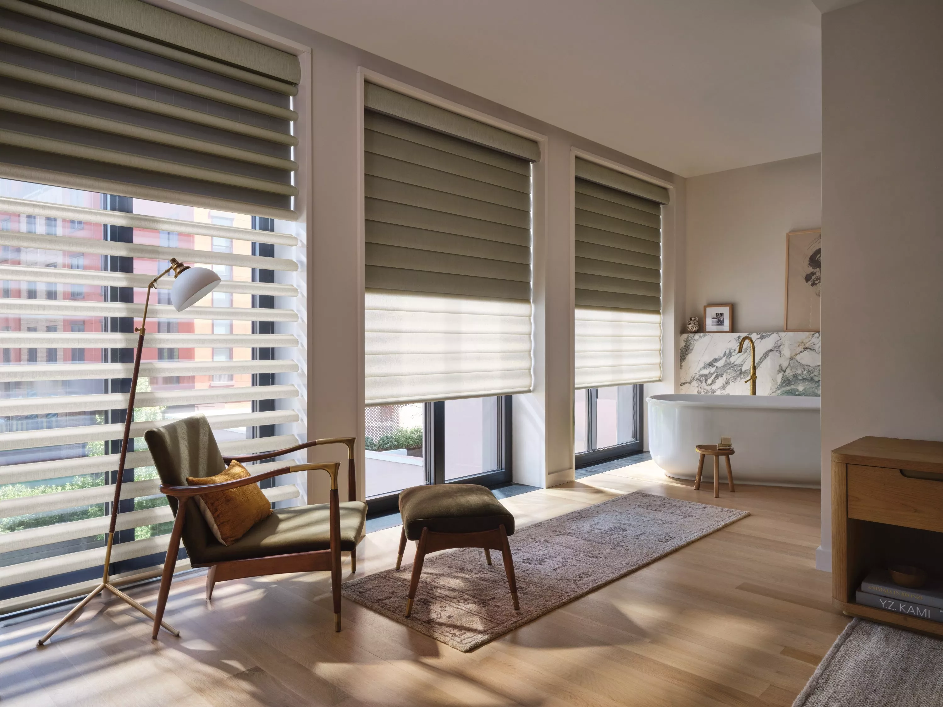 Modern living room with large windows, zebra roller blinds, armchair and ottoman, and a freestanding bath suite