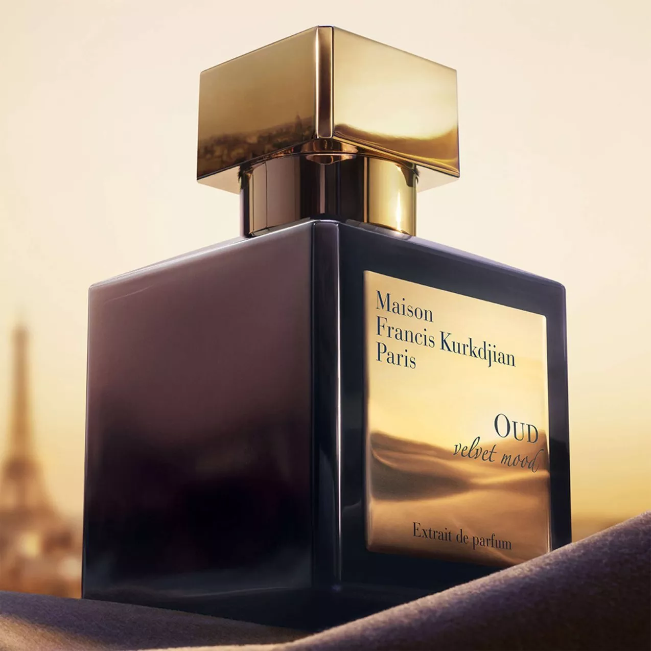 A bottle of Maison Francis Kurkdjian Paris OUD velvet mood Extrait de parfum with a metallic gold cap and label against a warm sunset backdrop showing a blurry Eiffel Tower in the distance.