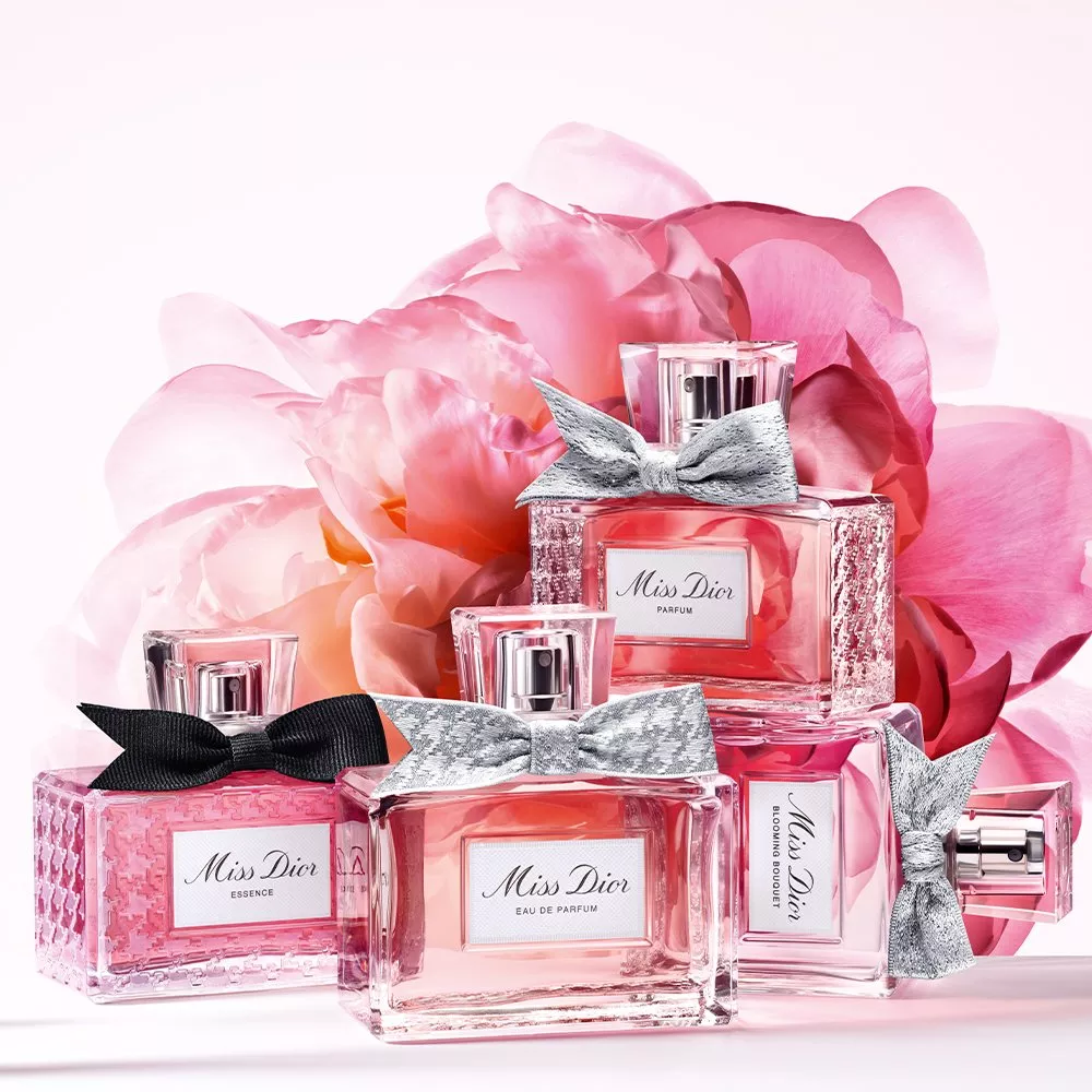 Miss Dior fragrance bottles in pink glass with silver bows, arranged before soft pink peony petals on white background