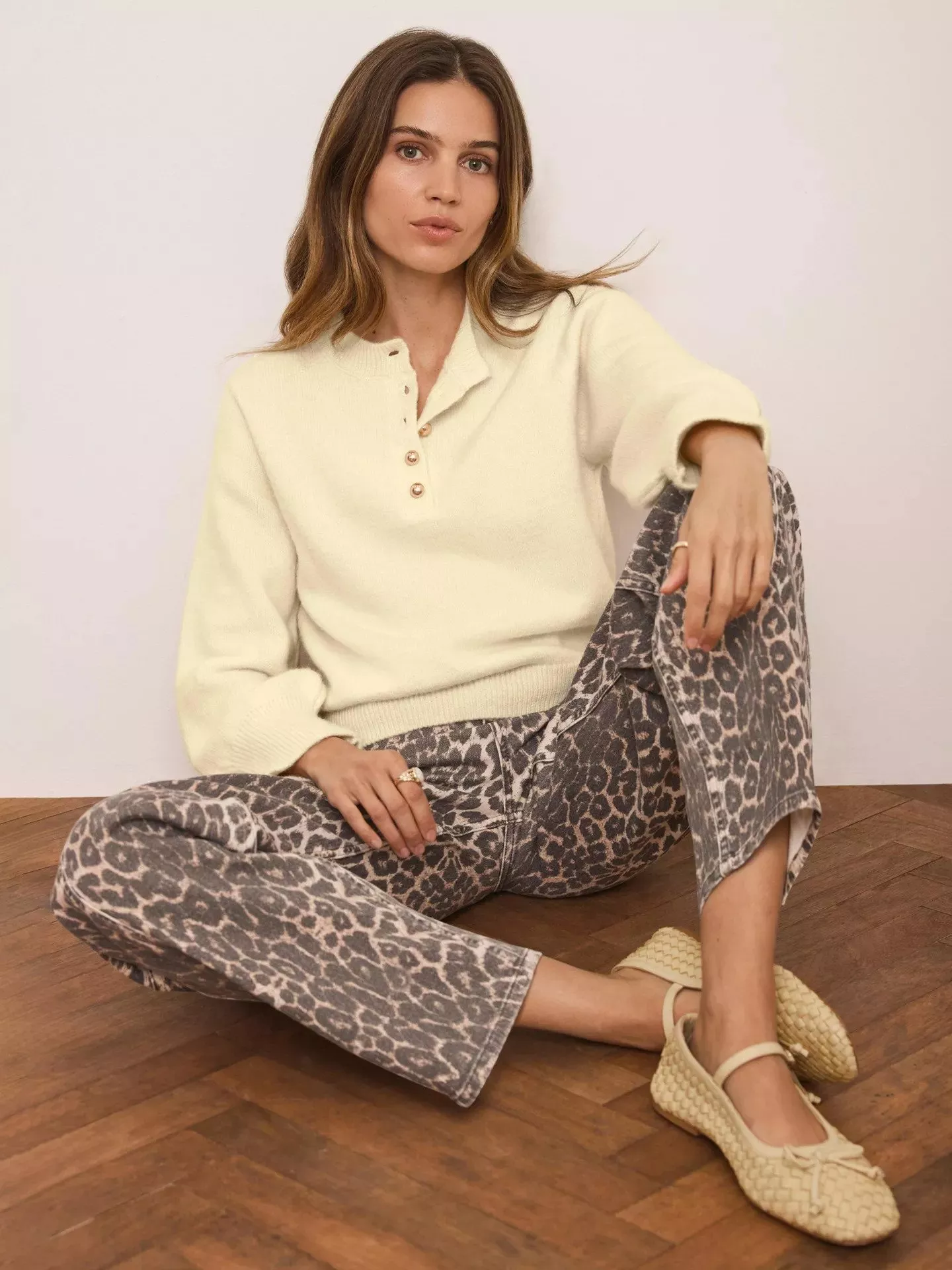 Woman in cream jumper and leopard print trousers, sitting on wooden floor, wearing beige woven shoes.