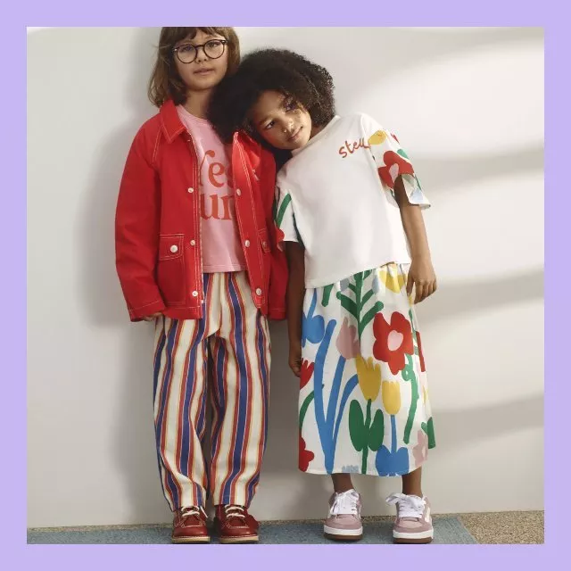 Two children in colourful outfits, one in red jacket and glasses, the other in floral dress, posing together indoors