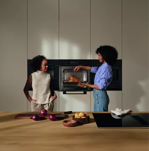 Modern kitchen scene with two women roasting chicken in built-in oven, fresh vegetables on wooden worktop