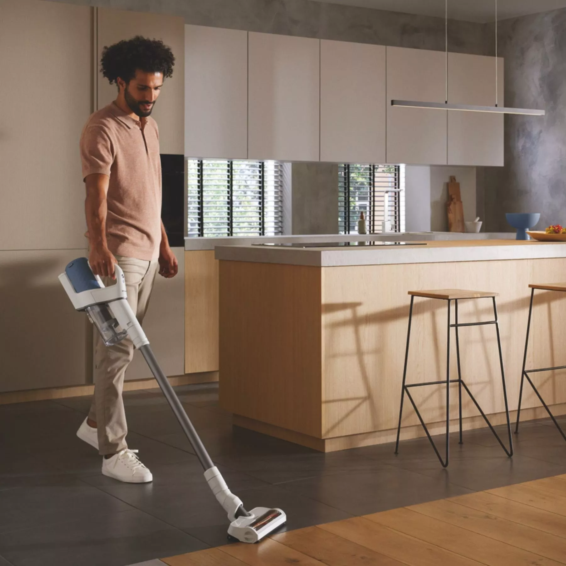 Man using cordless stick vacuum cleaner on tiled floor in modern kitchen, cleaning around island and stools