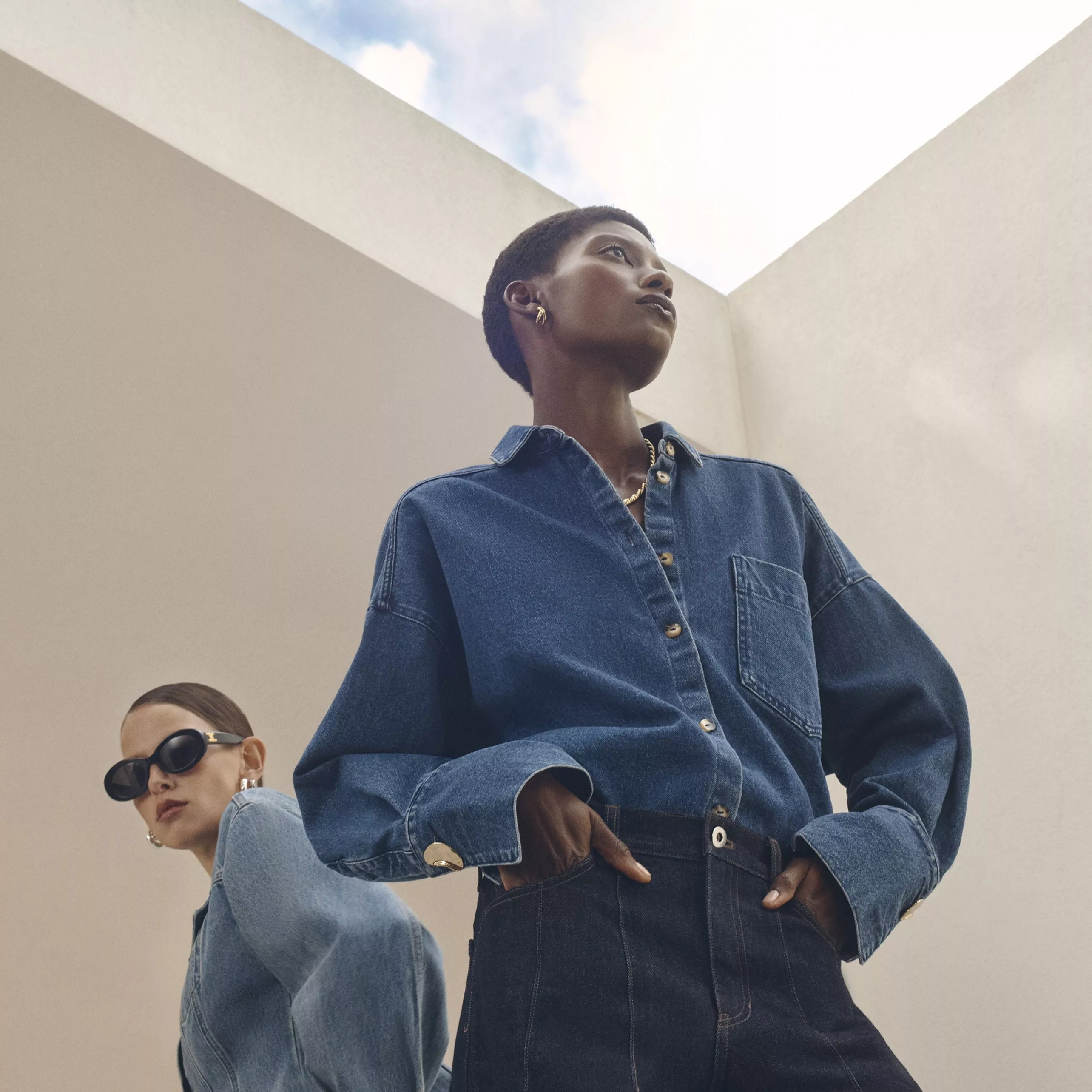 Two individuals in denim outfits pose against a white wall; one walks past, the other leans casually.