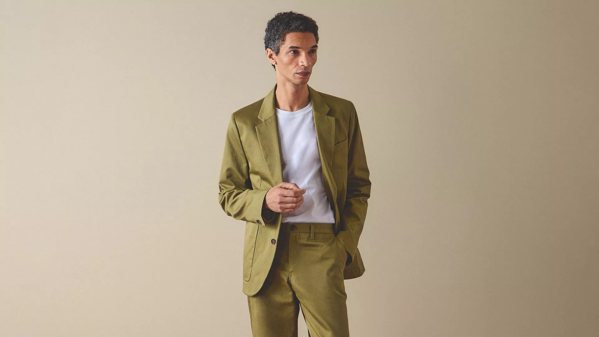 Male model wearing an olive green suit with white T-shirt, standing against a beige studio background, smart casual style