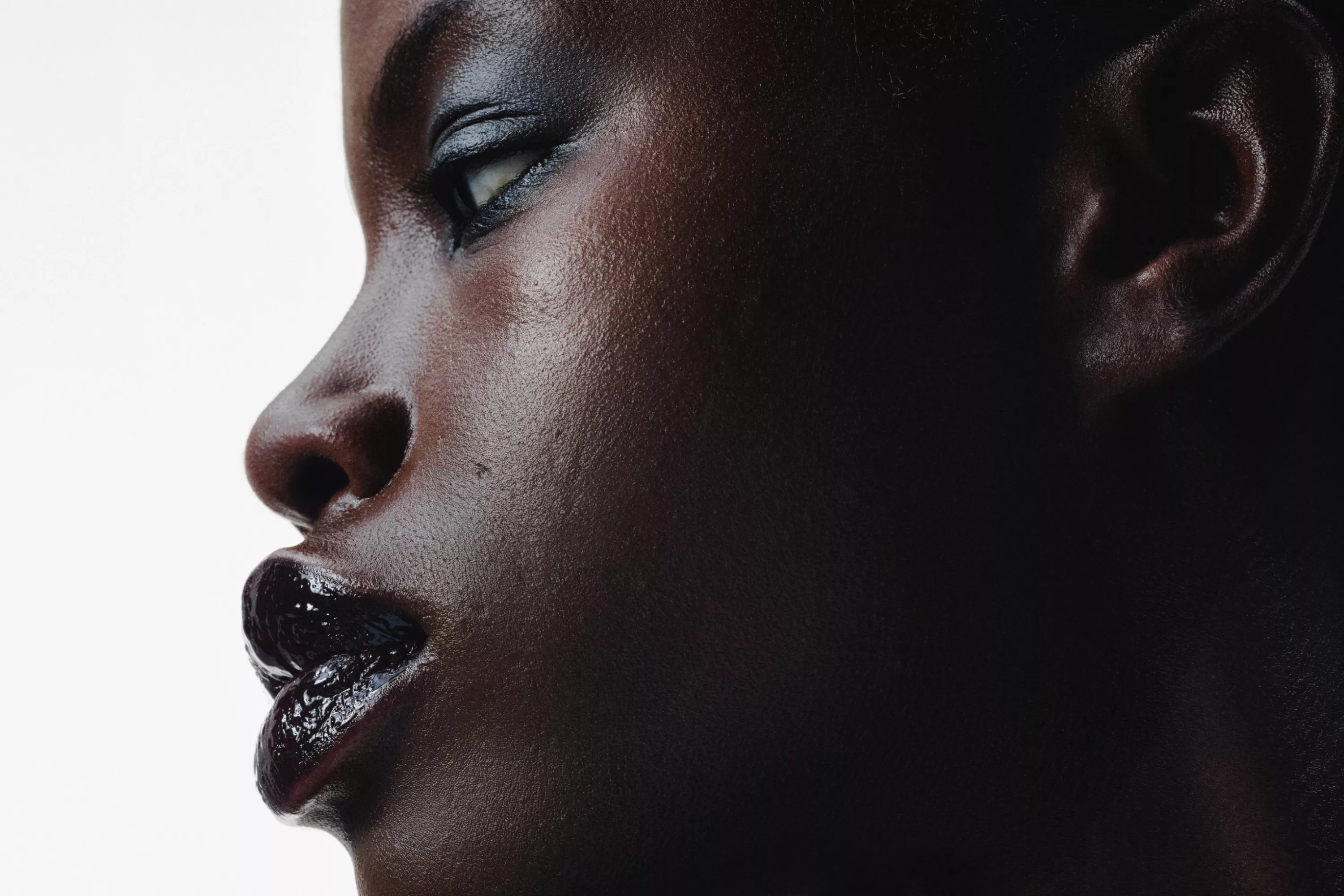 Close-up side profile portrait of a Black woman with glossy lips and smoky eye makeup on a white background