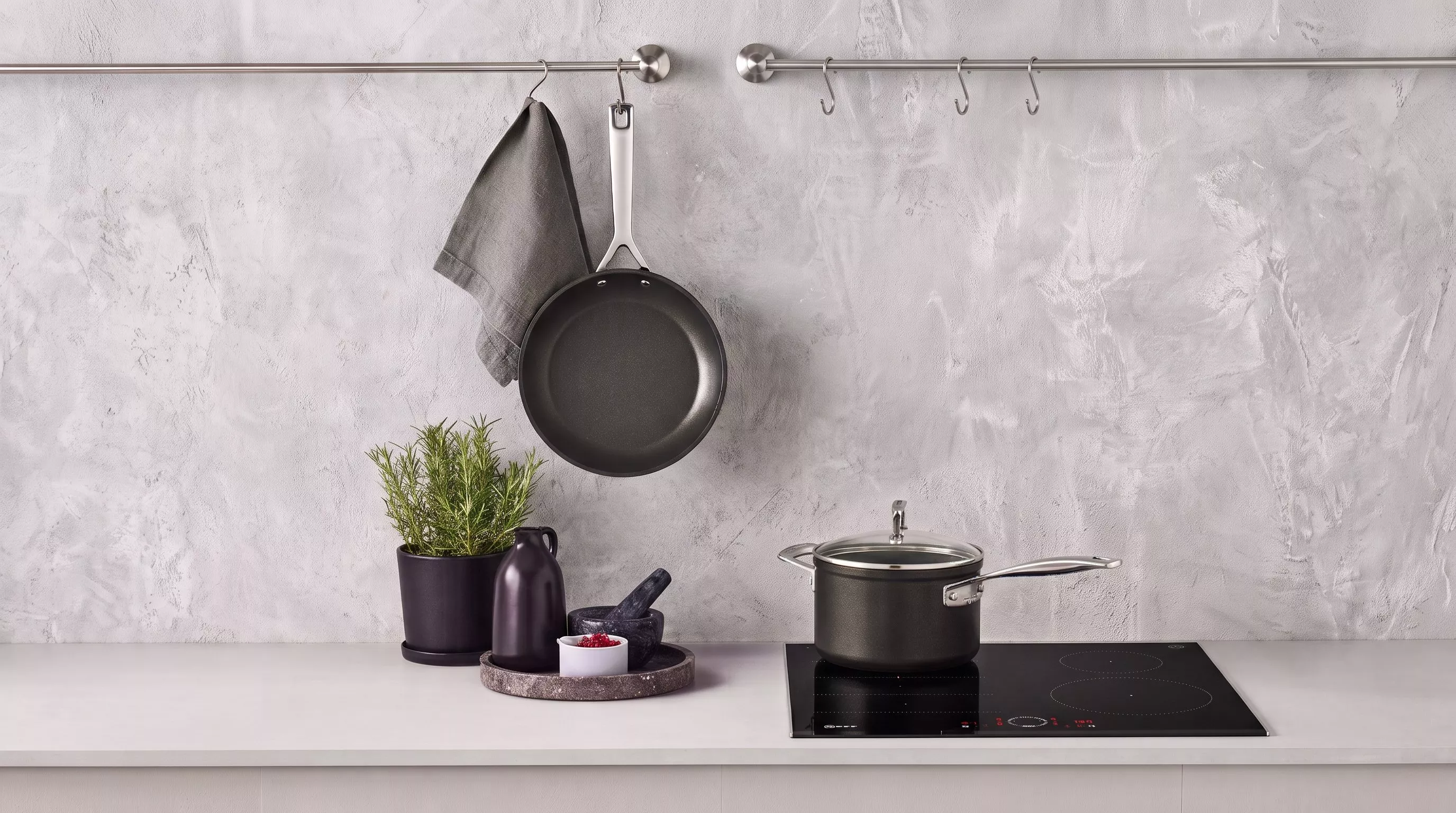 Black non-stick frying pan hanging on a grey wall next to a green potted plant and a black cooking pot on a gas hob.