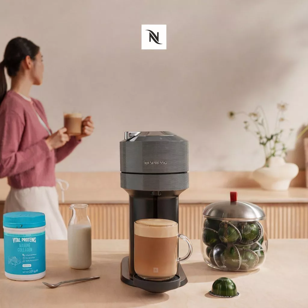 A person enjoys a coffee made by a sleek Nespresso machine on a wooden counter, surrounded by coffee pods.