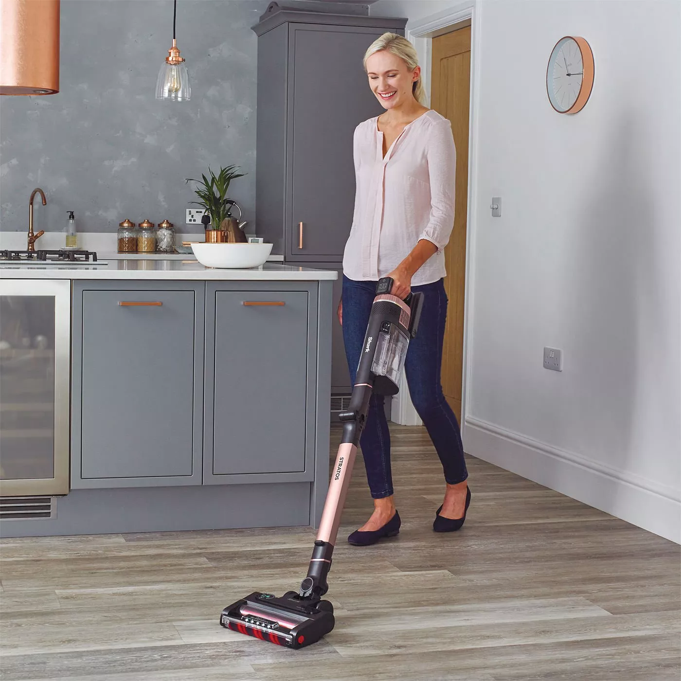 Woman using cordless stick vacuum cleaner on kitchen floor in modern grey kitchen with copper accents
