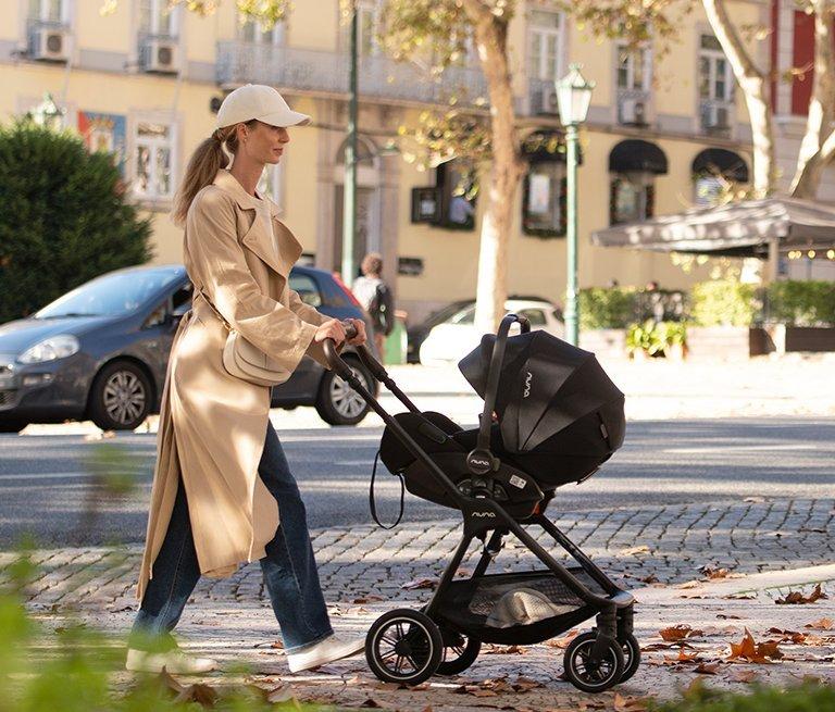 Side view of a person in a tan trench coat and cap pushing a black Nuna stroller down a sunlit city sidewalk with autumn leaves and parked cars in the background.