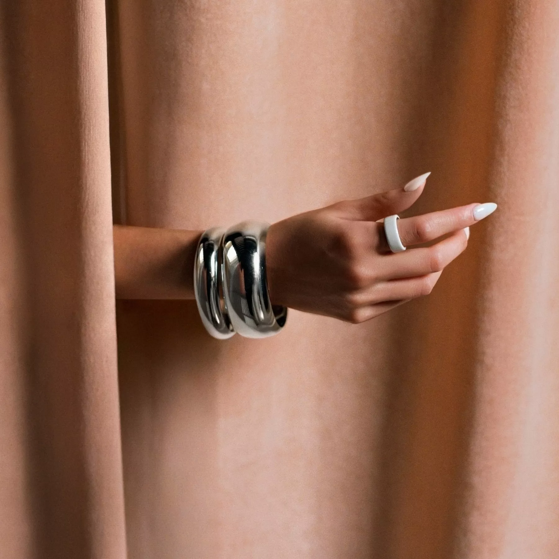 A woman's hand extends from behind a draped peach curtain, showcasing a white smart ring on her index finger and two chunky, polished silver bangle bracelets on her wrist.