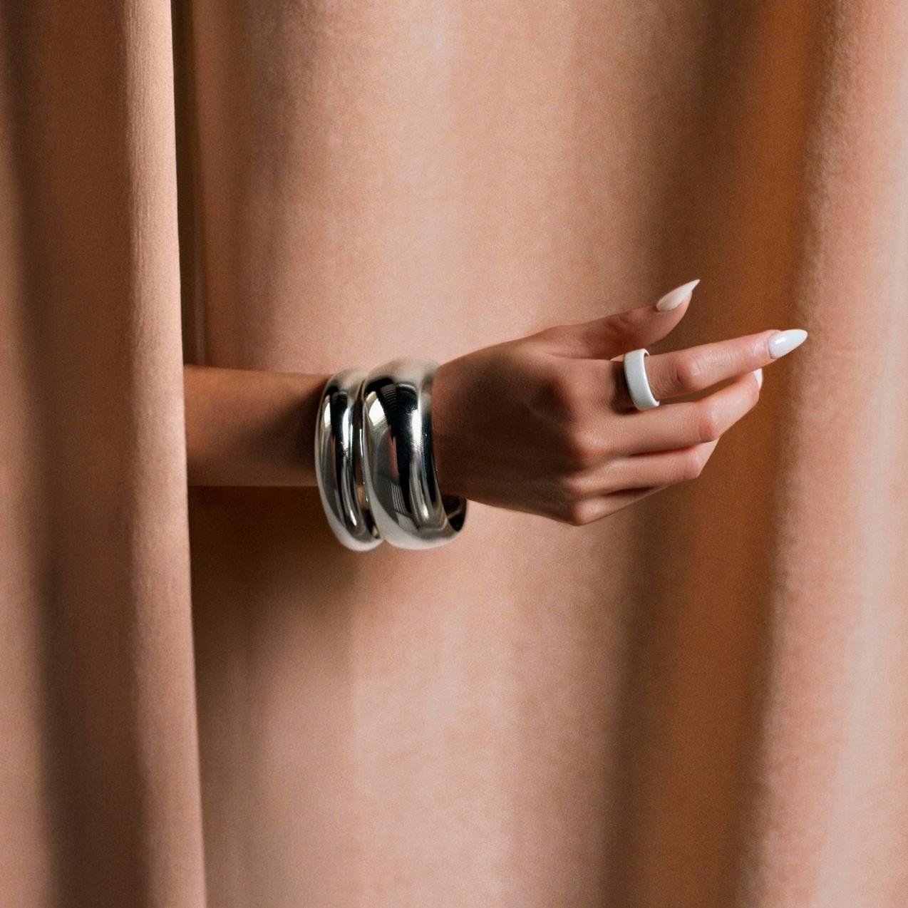 A woman's hand extends from behind a draped peach curtain, showcasing a white smart ring on her index finger and two chunky, polished silver bangle bracelets on her wrist.