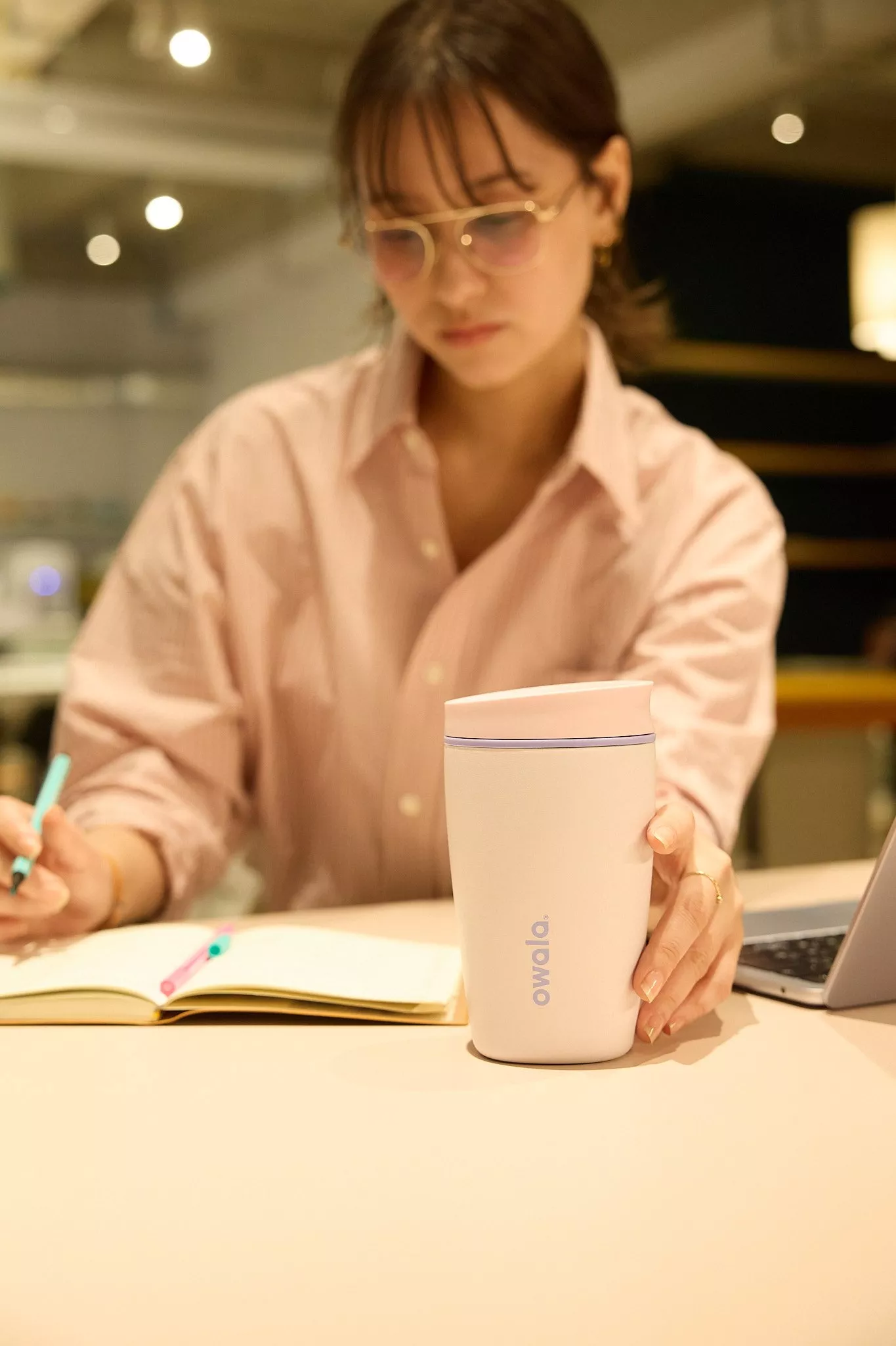 Woman studying at desk with laptop and notebook, holding a white Owala insulated travel tumbler in café