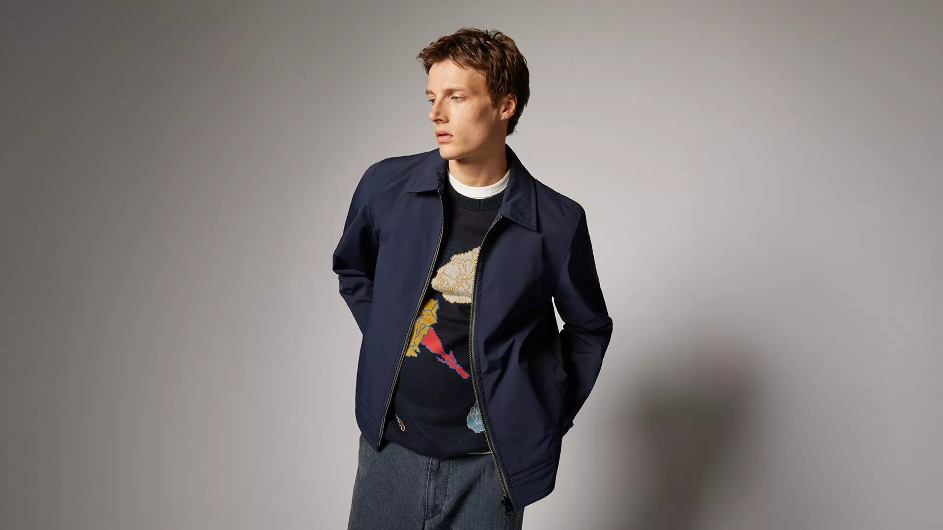 A person wearing a dark jacket over a patterned sweater, standing against a plain grey background.