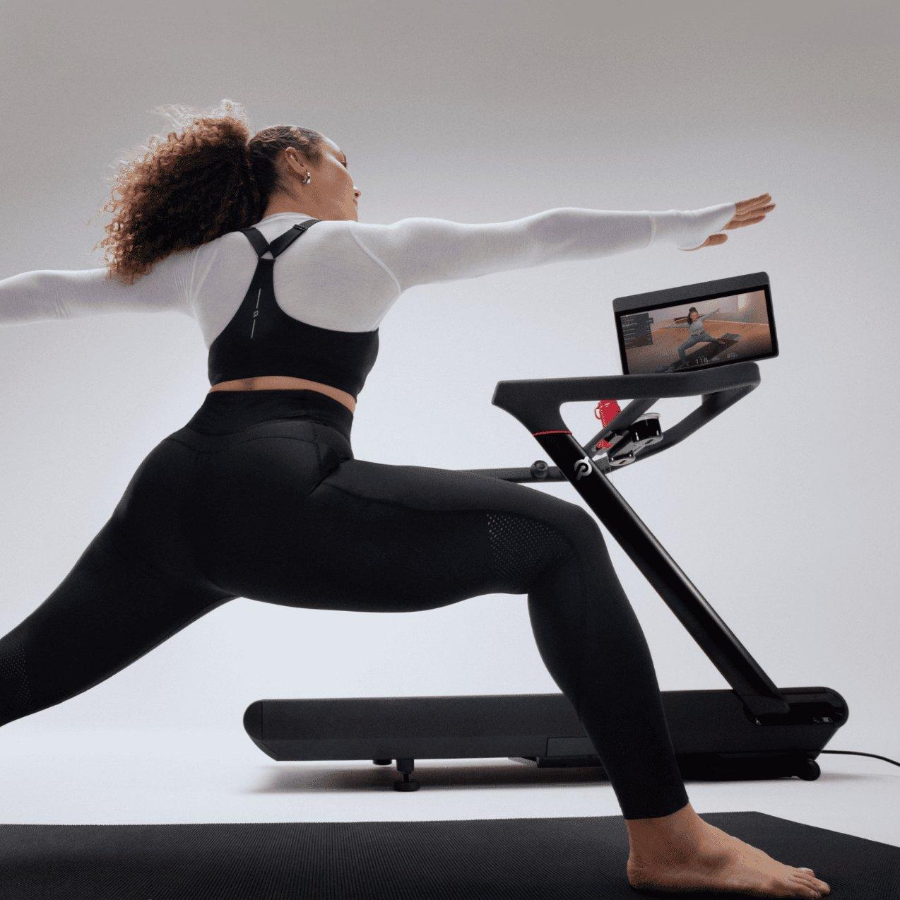 Woman doing yoga warrior pose beside Peloton treadmill, following on-screen fitness class at home studio