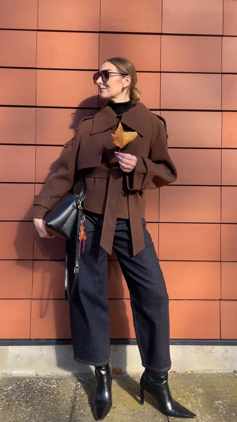 Stylist wearing a brown jacket, black roll neck jumper, denim jeans, black heeled boots and carrying a black bag