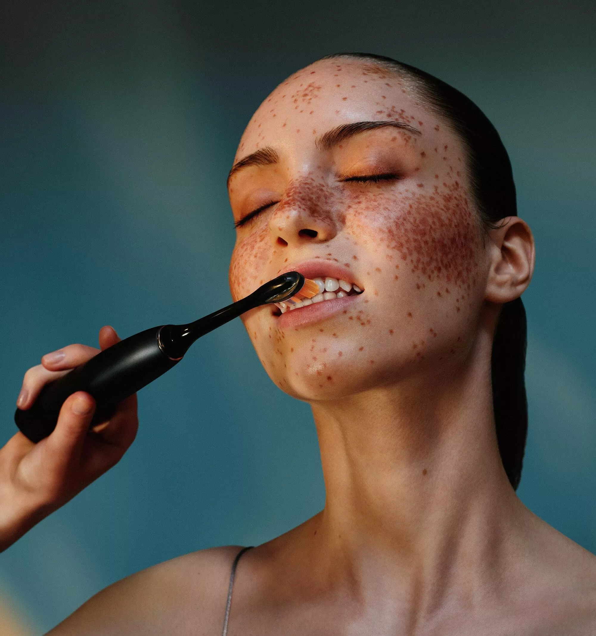 A person with freckles uses an electric toothbrush, eyes closed, against a soft blue background.