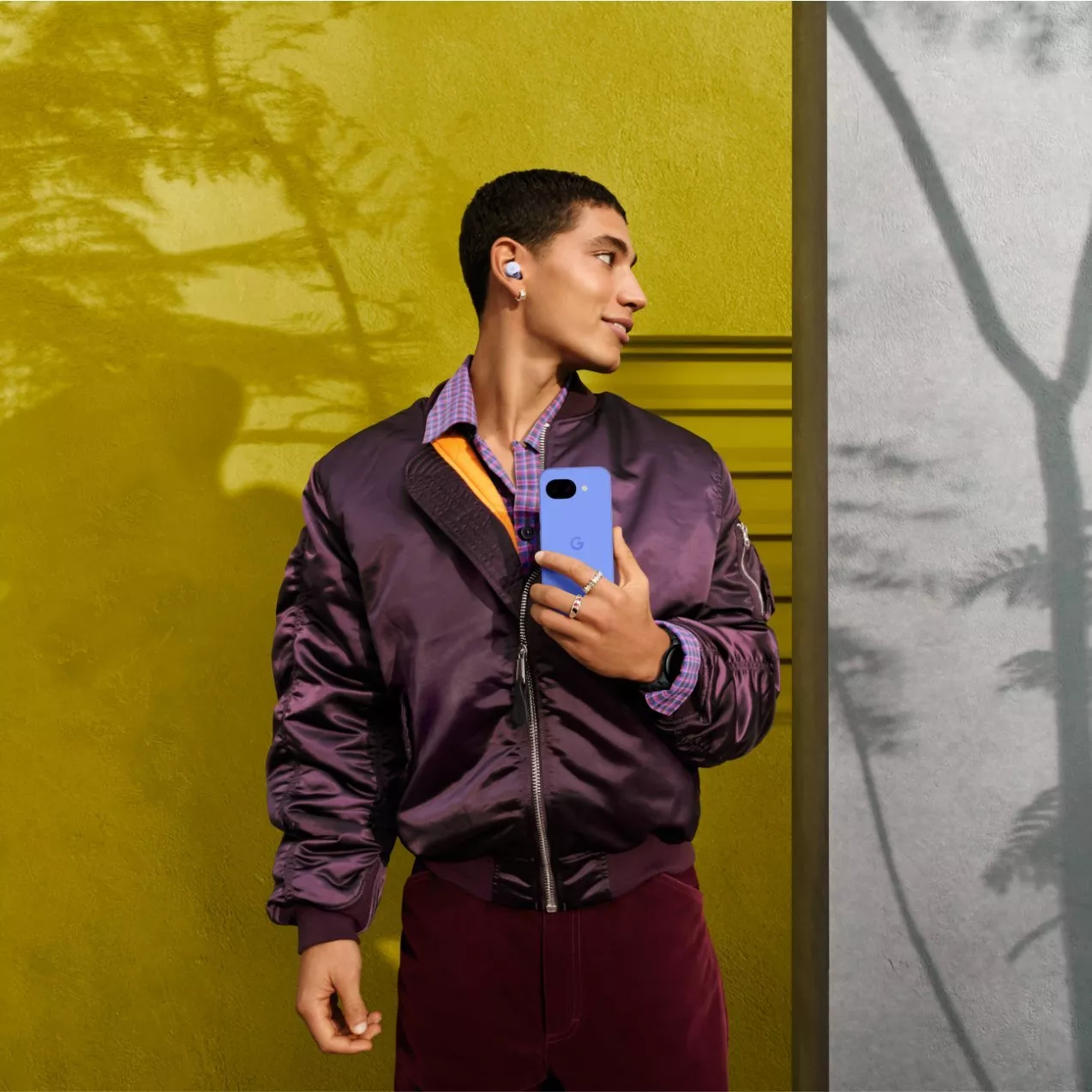 Stylish man holding a blue Google Pixel smartphone, wearing wireless earbuds, standing by a yellow wall outdoors