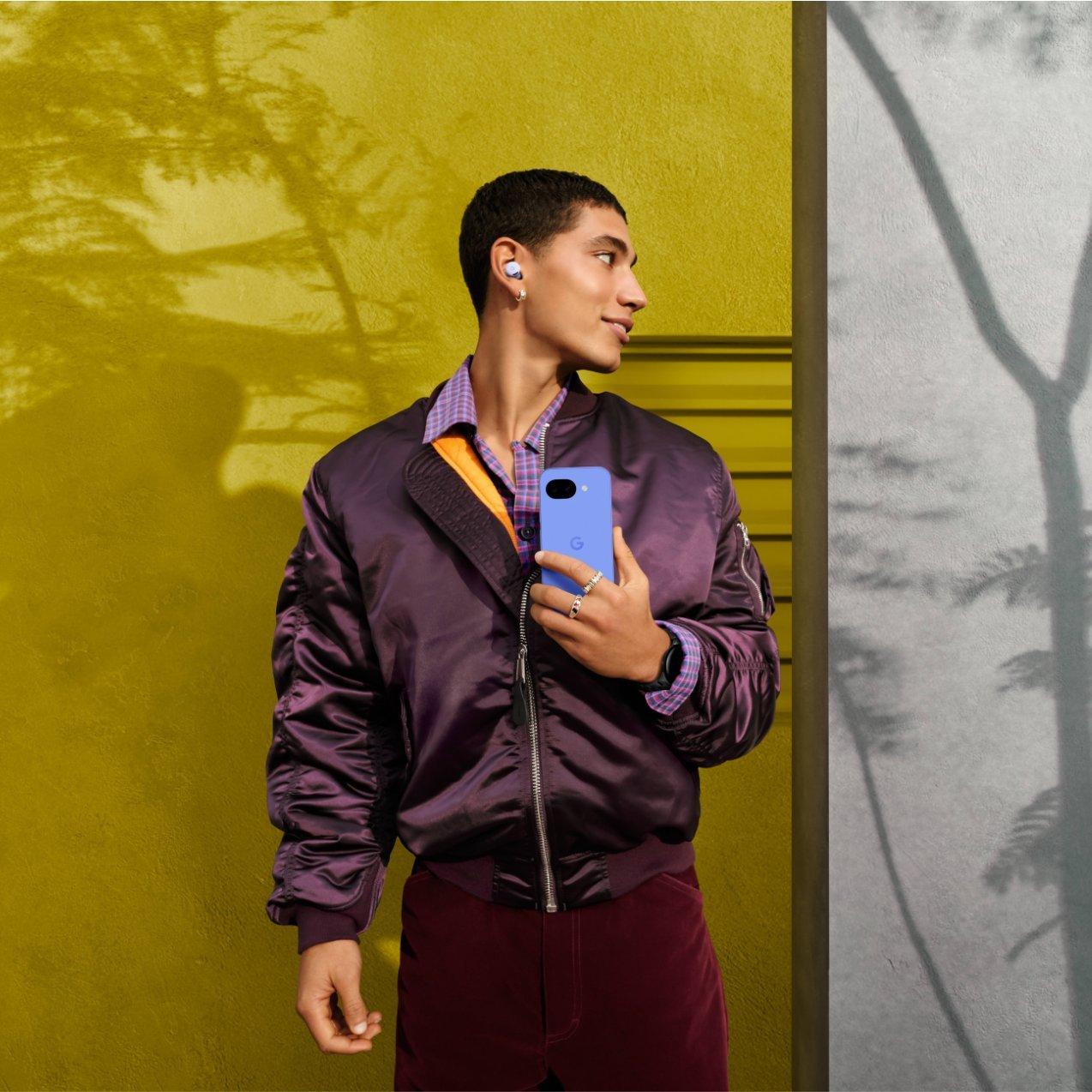 A man wearing a purple bomber jacket holds a blue Google Pixel phone against a vibrant yellow and grey wall with tree shadows.