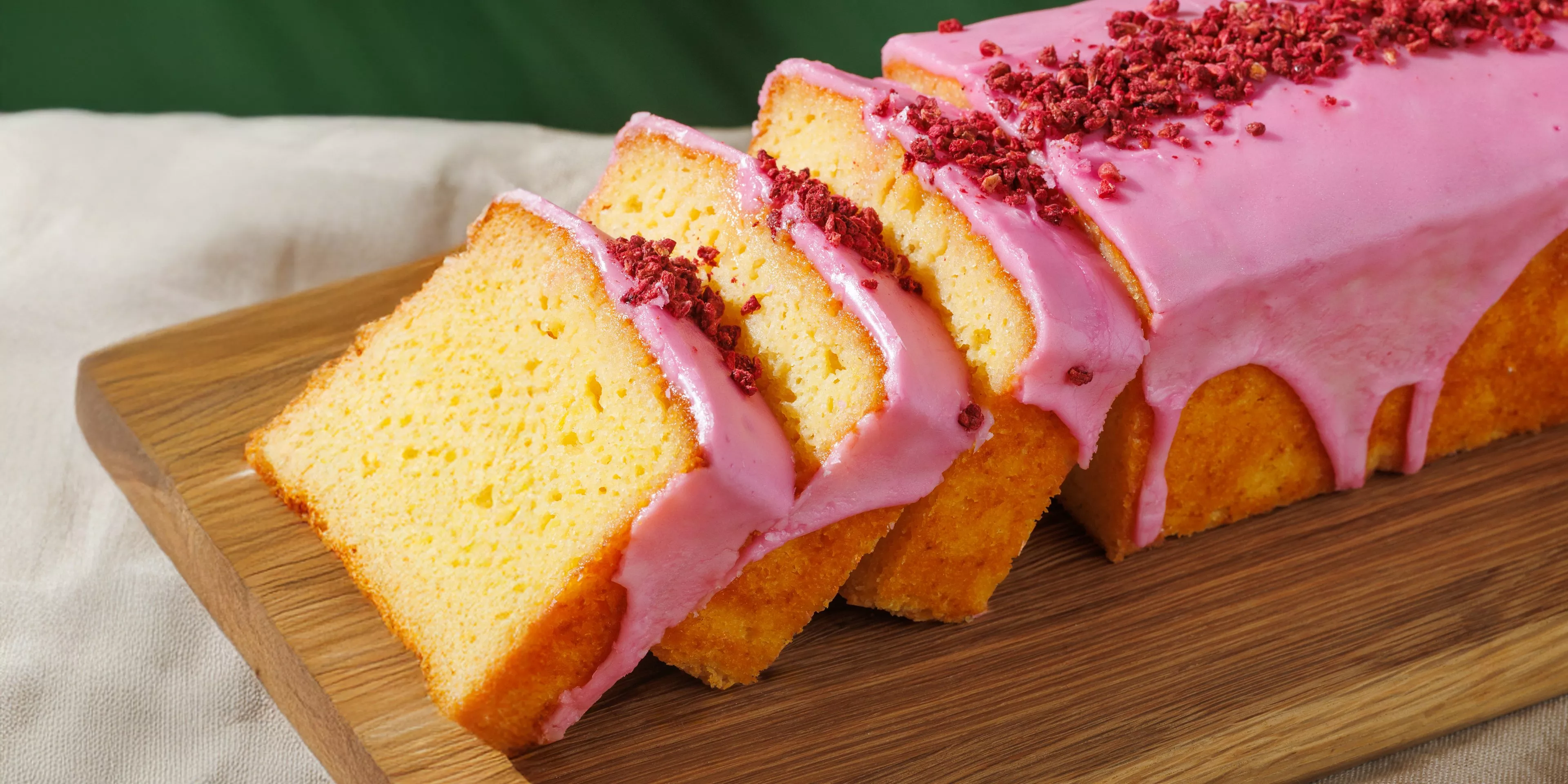 Sliced lemon drizzle loaf cake with pink icing and freeze-dried raspberry topping on a wooden board